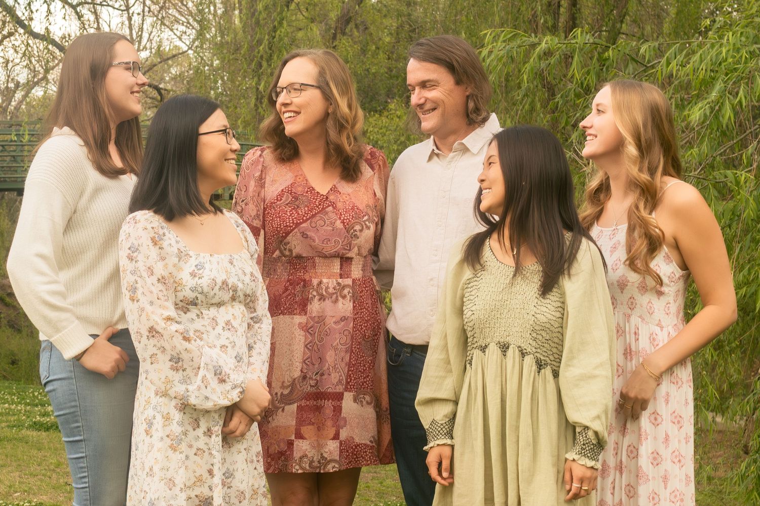 A family wearing flowy bohemian dresses and casual attire stand together outdoors under a willow tree during a family portrait session in FW