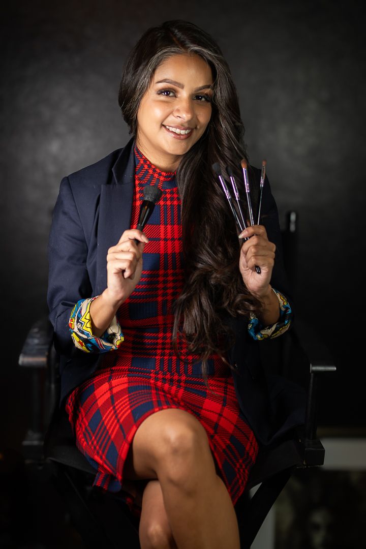A makeup artist in a plaid dress and navy blazer holds brushes while sitting in a professional setting.