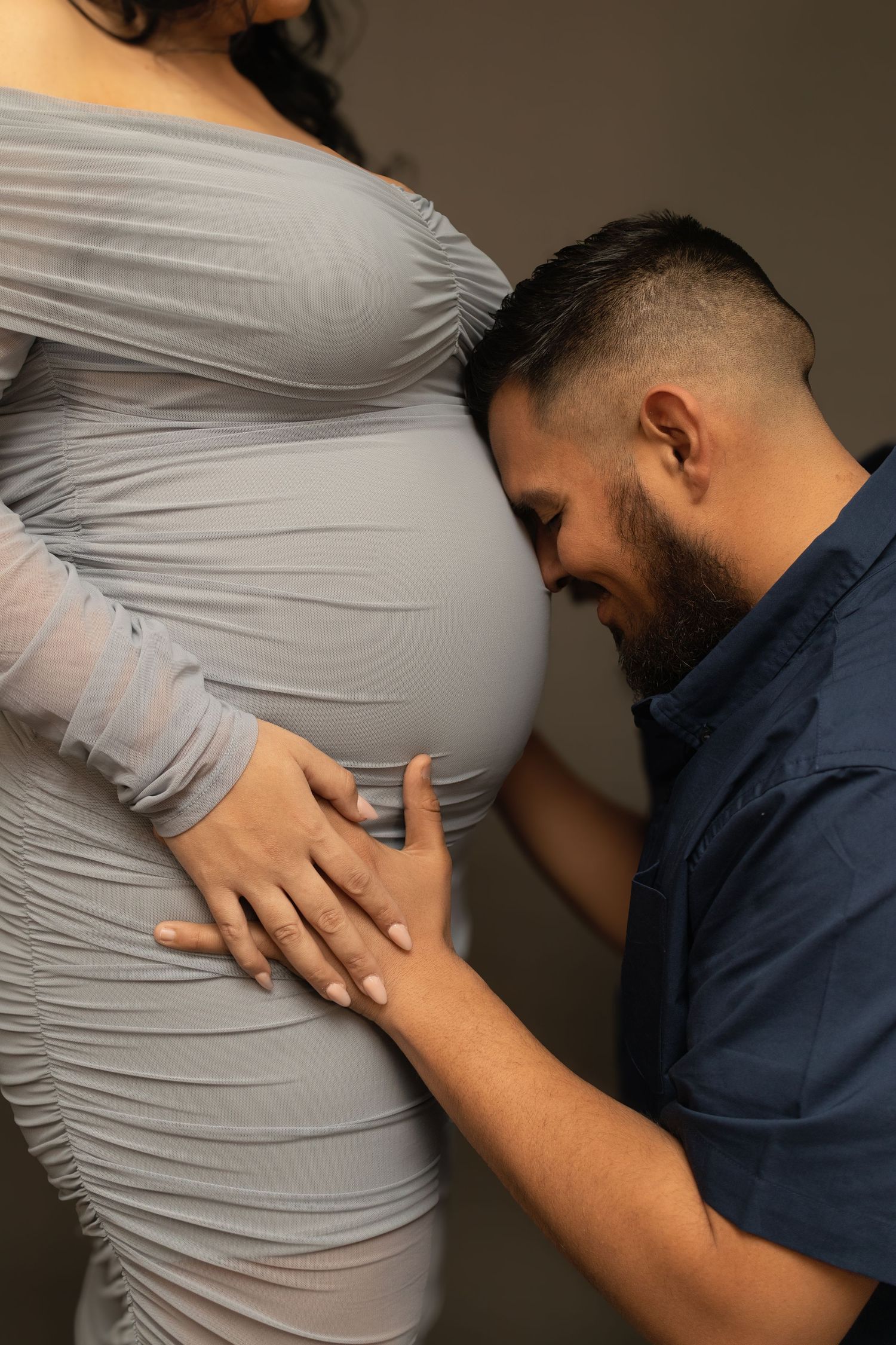 A man smiles against his wife's expectant belly during a studio maternity portrait session in the DFW metroplex.