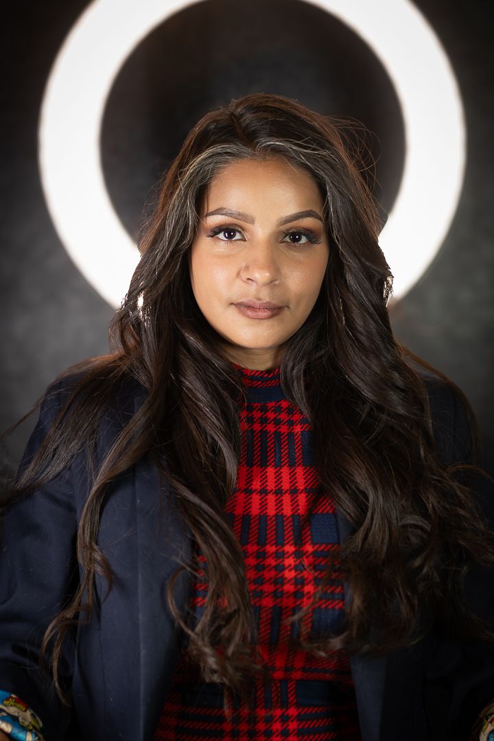 A portrait with a ring light shows a person in a plaid top and navy blazer.
