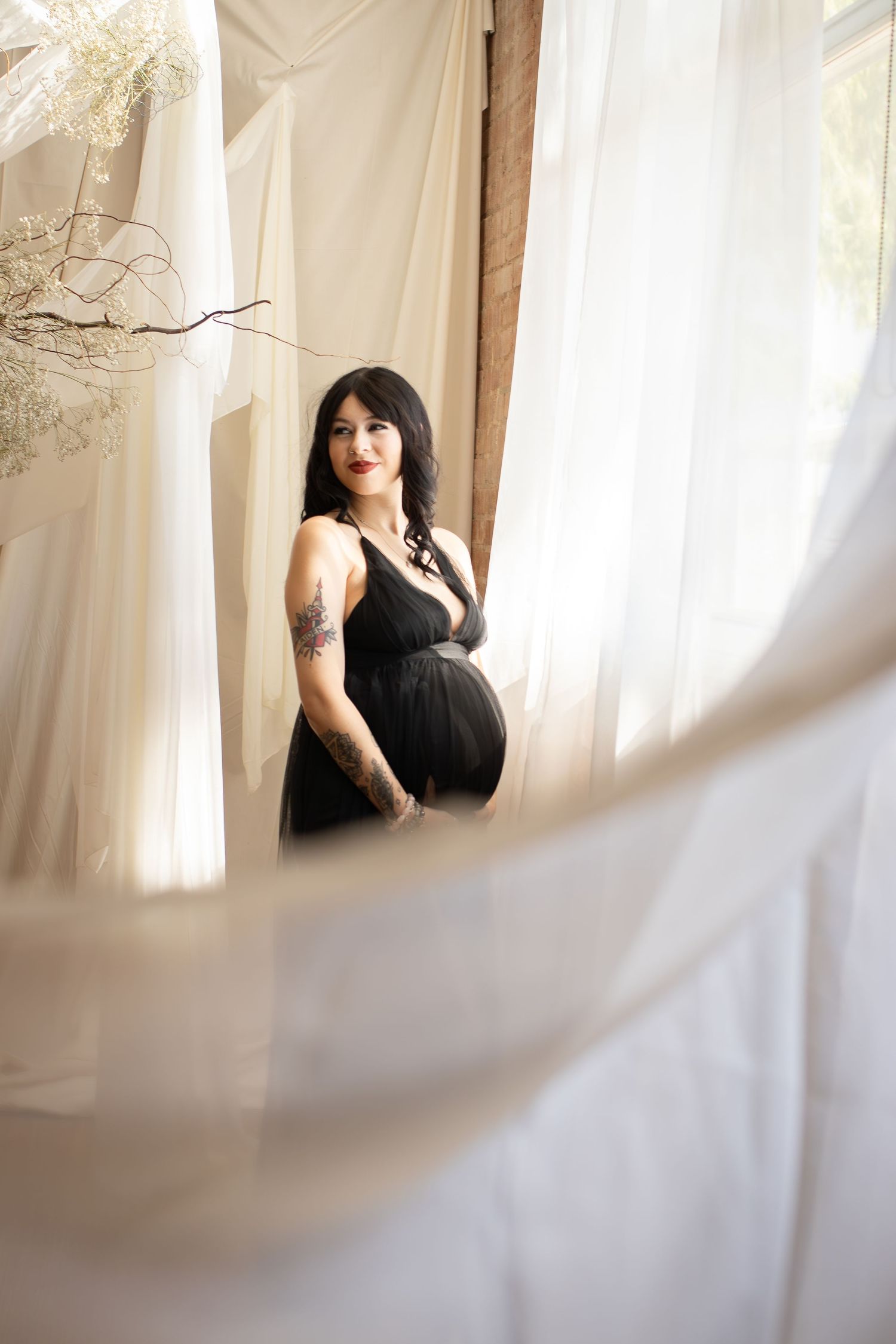 A maternity portrait of an expectant mother in a black dress near sheer curtains.