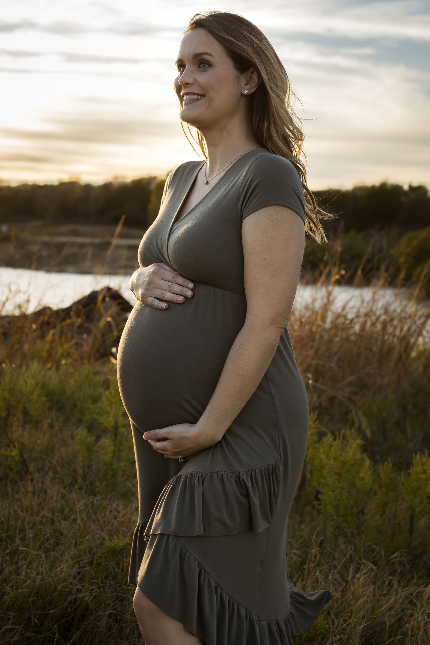 Expectant mother in gray dress smiling during sunset maternity photo session.