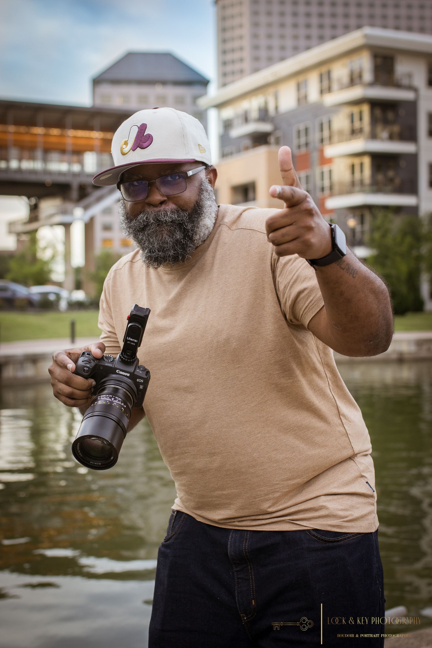 Happy photographer with beard giving thumbs up while holding camera near waterfront.