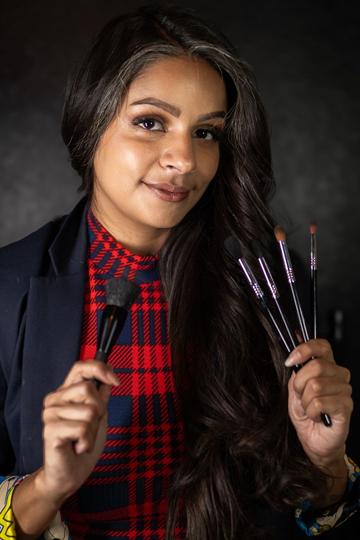 Professional makeup artist wearing a red plaid top and navy blazer holds up various cosmetic brushes against dark background.