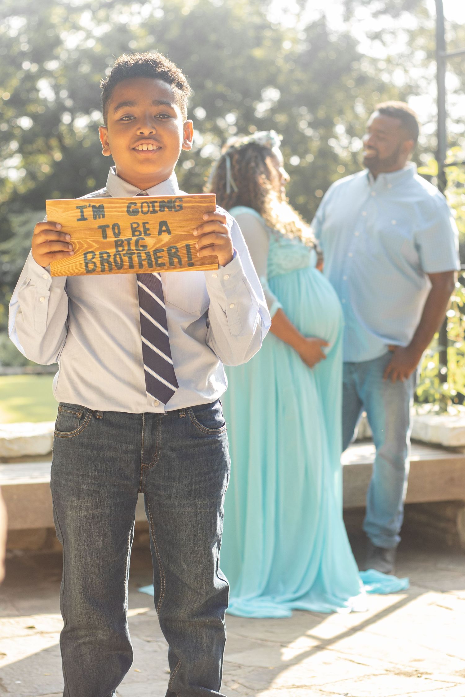 Child holding 'Going to be a Big Brother' sign with parents in background during maternity photoshoot.
