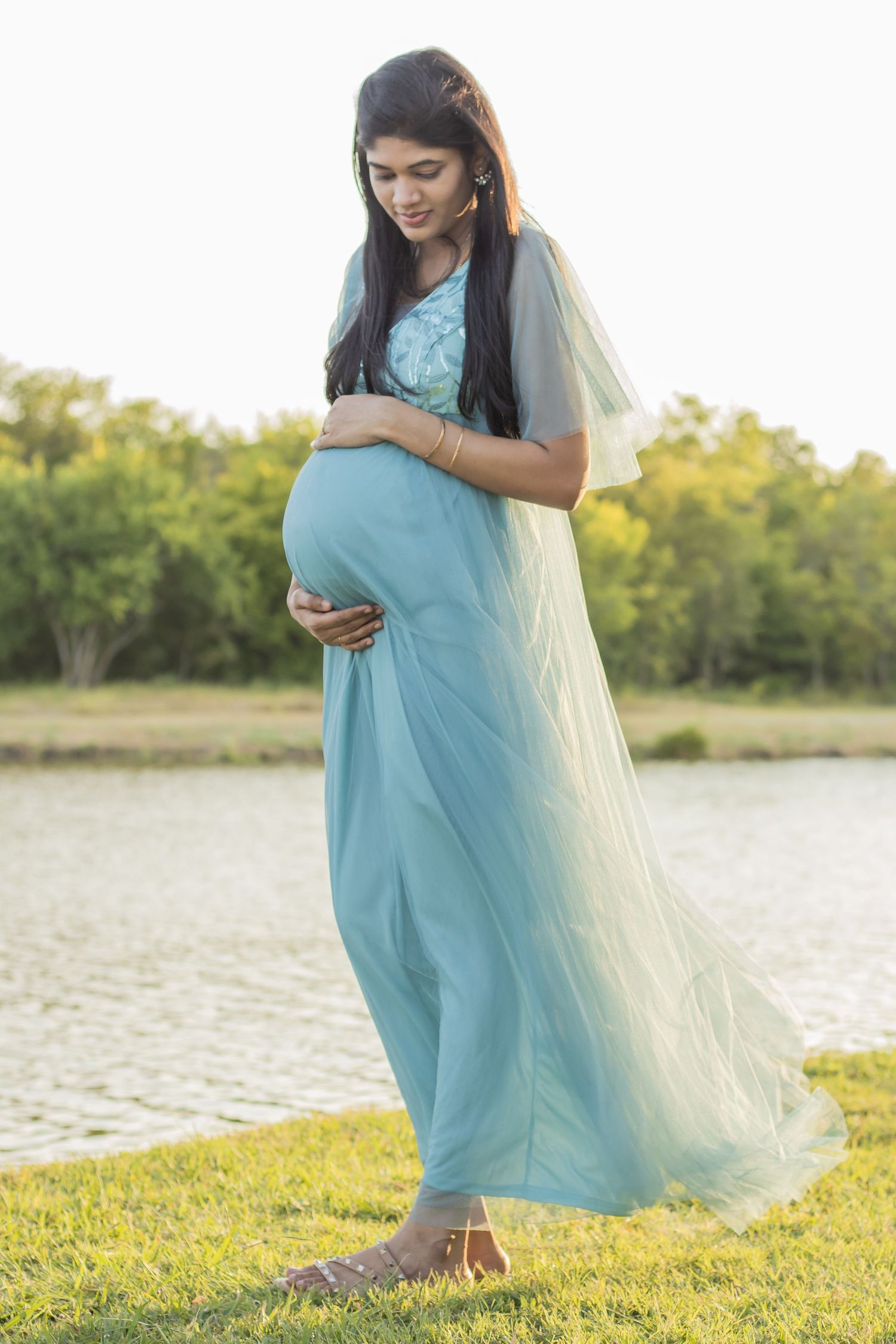Maternity photo of expecting mother in flowing blue dress and sheer overlay by lake at sunset.