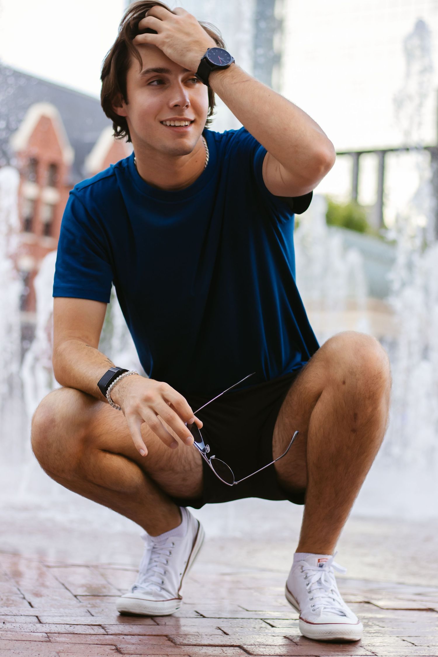 Casual portrait of person in blue shirt squatting on brick pavement with hand on head.
