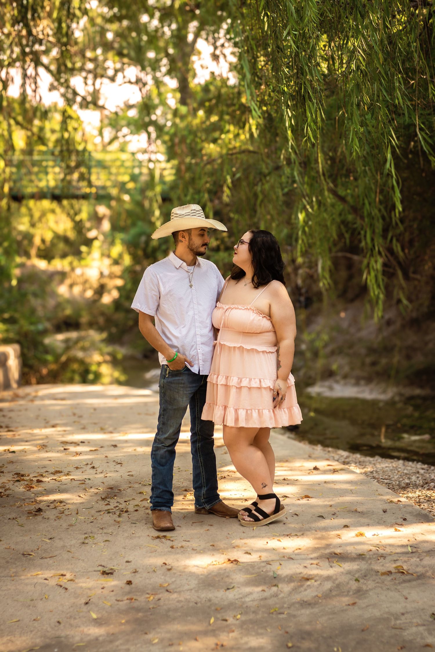 A romantic couple stands together on a sunlit path under weeping willow trees in a soft summer glow.
