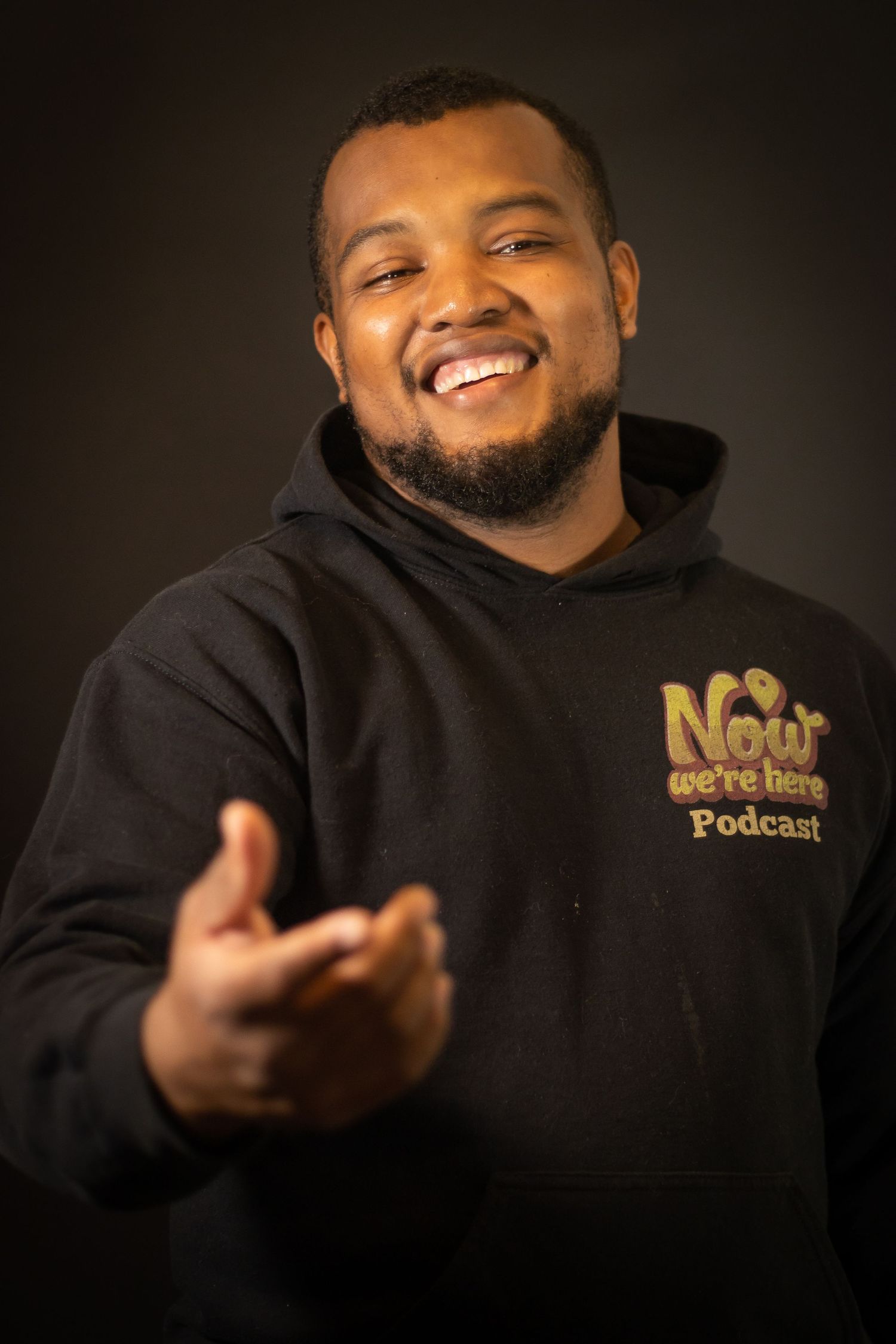 A person wearing a black hoodie with 'Now We're Here Podcast' logo smiles warmly at the camera.
