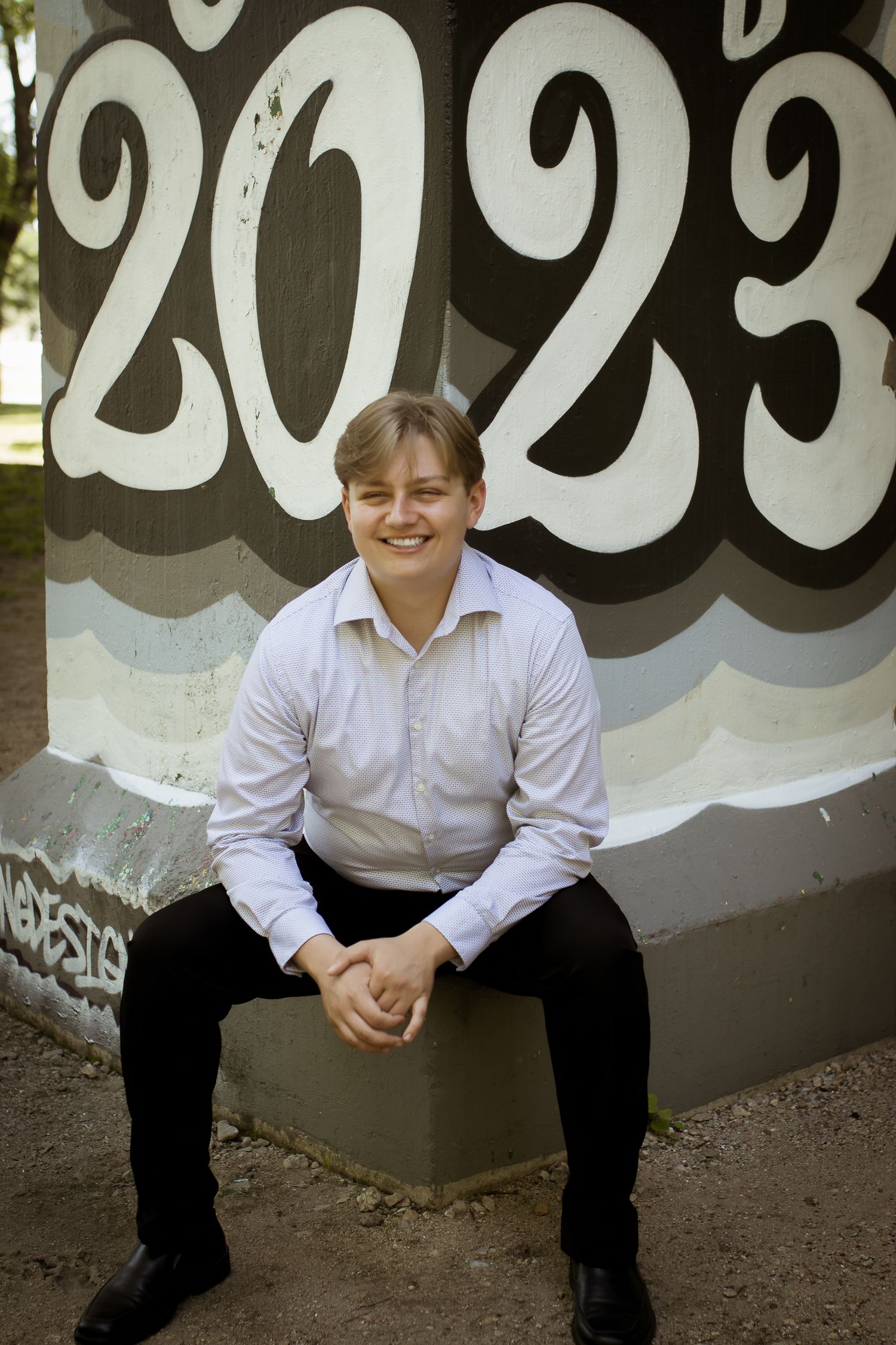 A graduate in formal attire sits on a bench in front of a wall with large painted numbers reading 2023 during a graduate portrait session.