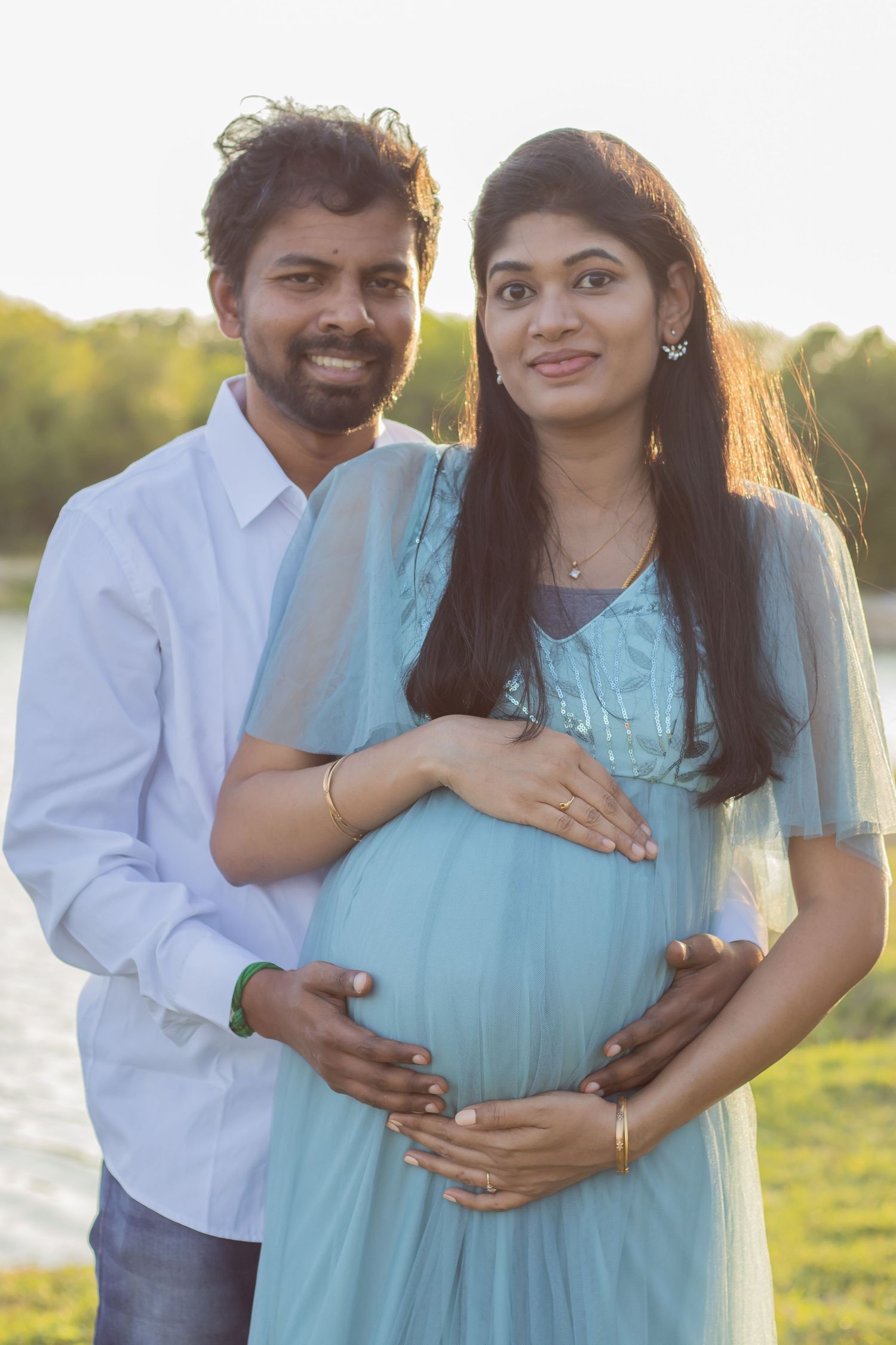 Maternity portrait of couple in blue dress and white shirt embracing outdoors at sunset.