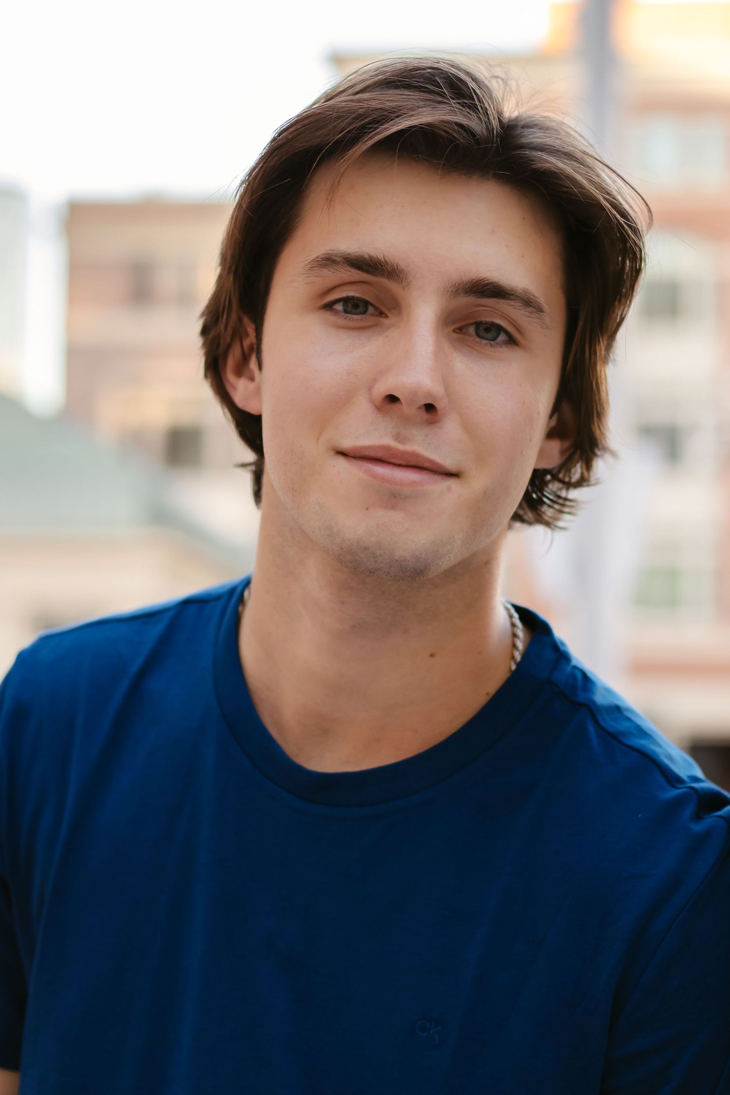 A person wearing a navy blue t-shirt smiles warmly at the camera in an outdoor setting with blurred buildings behind them in Fort Worth, TX.
