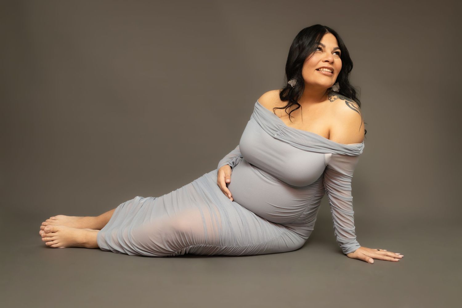 A maternity portrait features a woman in a fitted grey dress posed elegantly against a dark backdrop.