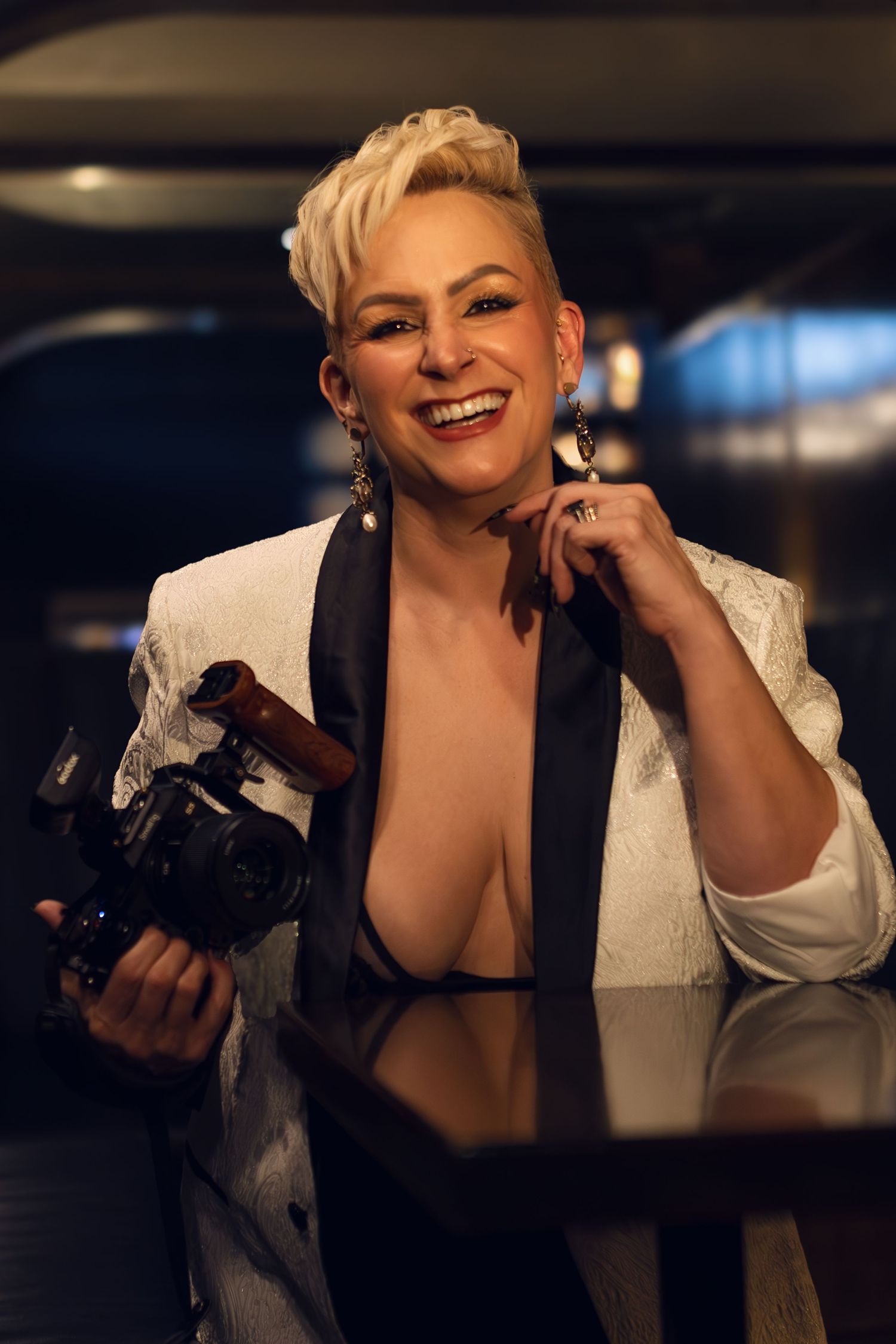 Professional photographer in white blazer holding camera and smiling in studio setting.