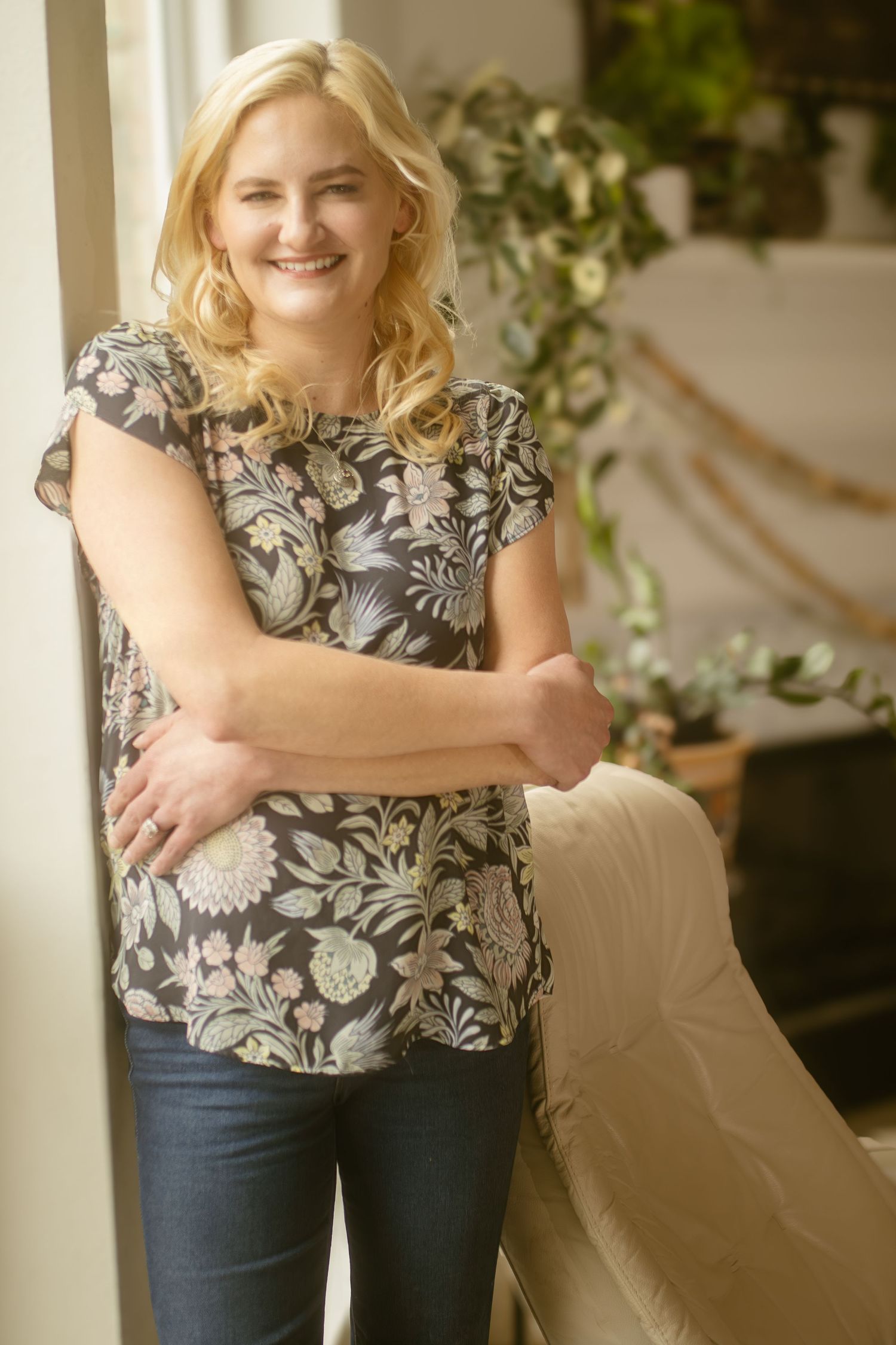 Person wearing floral print cap sleeve blouse and jeans poses by window with potted plants in background for a DFW branding session.
