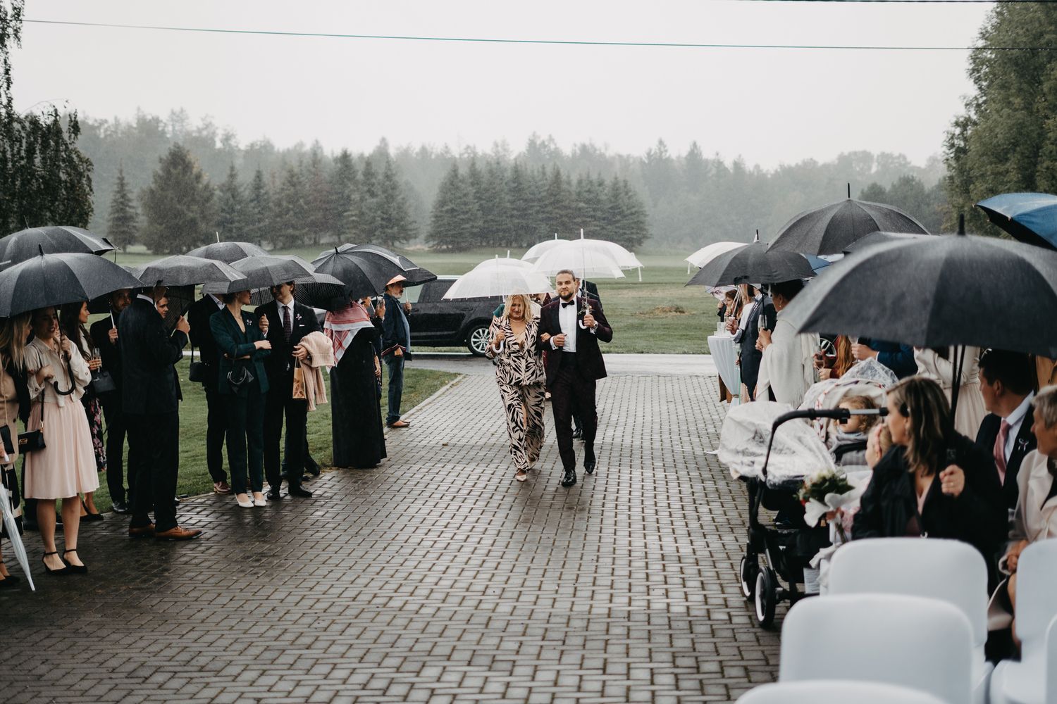 Wedding guests holding black umbrellas line a pathway on a rainy day at an outdoor ceremony.