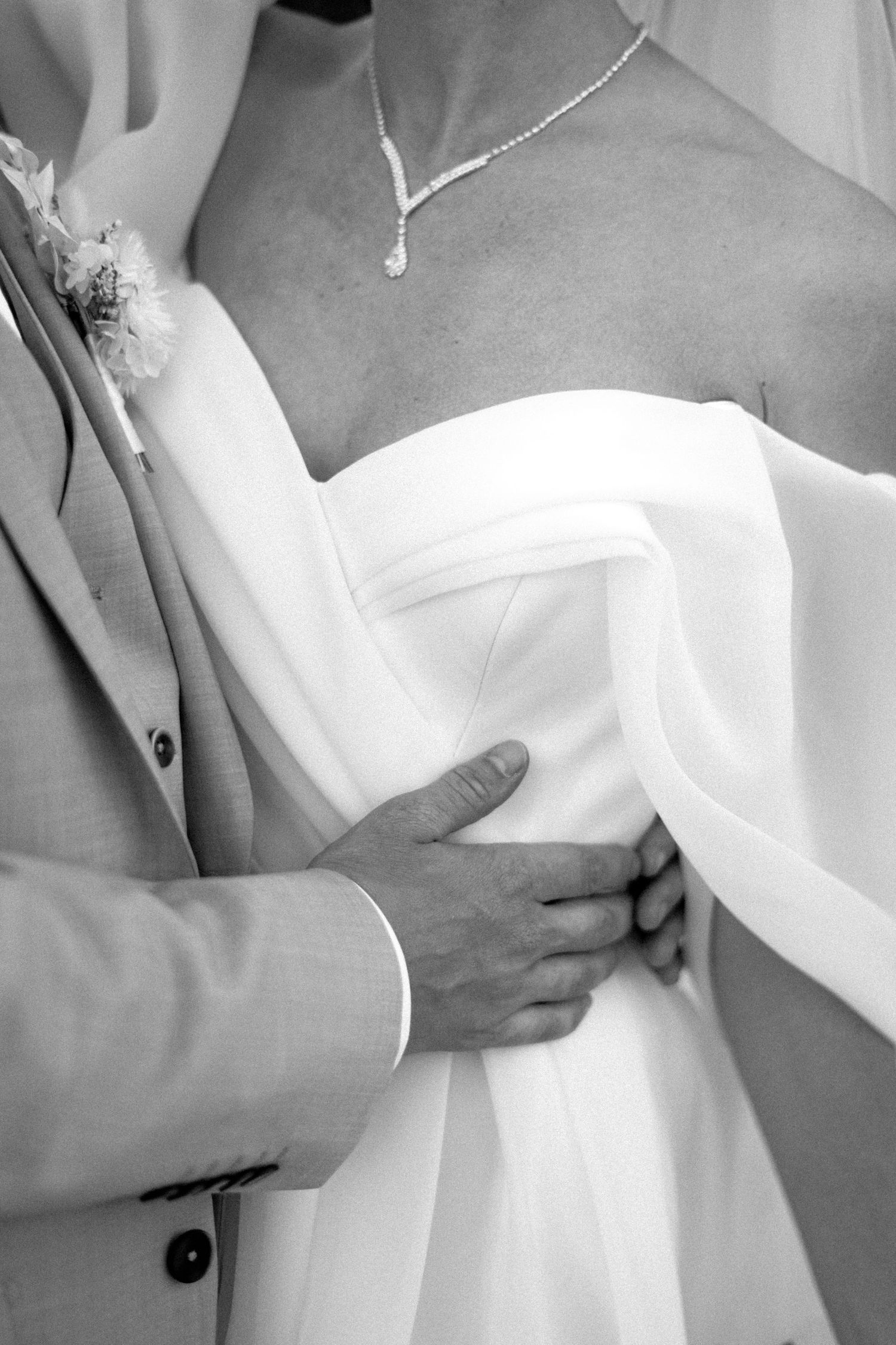Close-up black and white photo of a traditional wedding dress with a delicate necklace and hand gently touching the fabric.