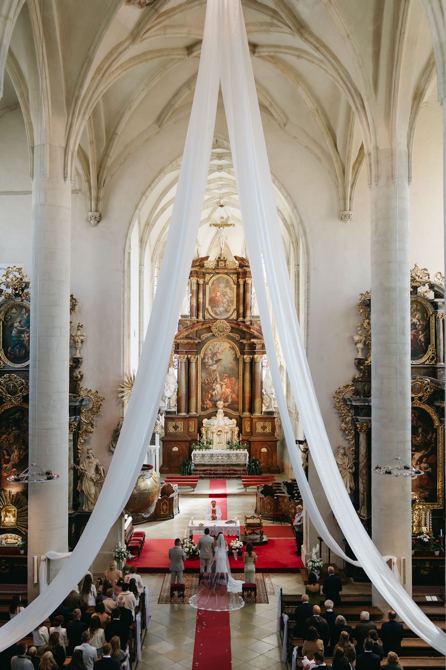 Elegant church wedding ceremony with white draping, ornate baroque altar, red carpet aisle, and guests seated in wooden pews.