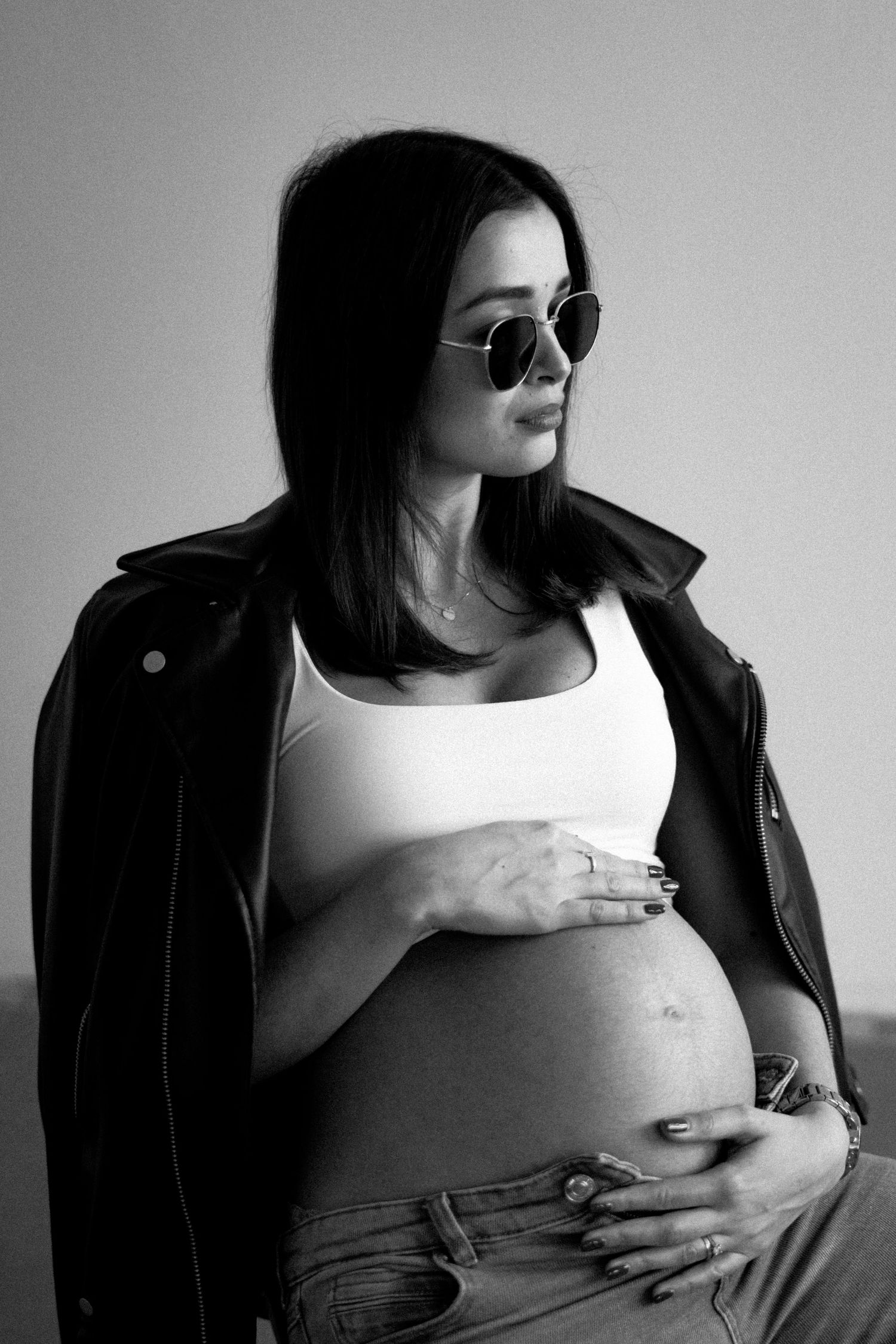 Black and white maternity photo of pregnant woman in leather jacket and sunglasses touching her belly.