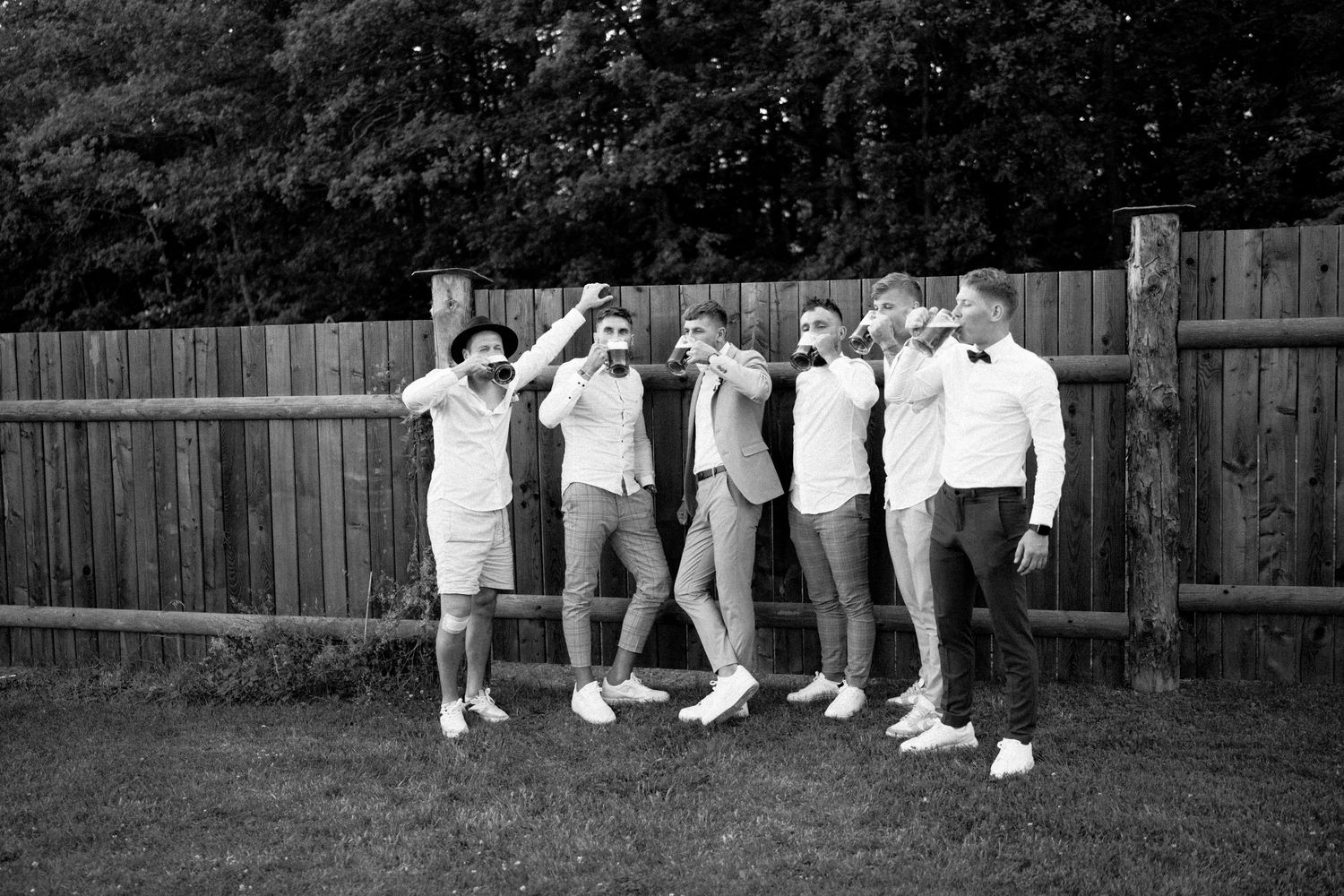 A black and white photo shows a group of people posing playfully in casual attire against a wooden fence in a backyard.