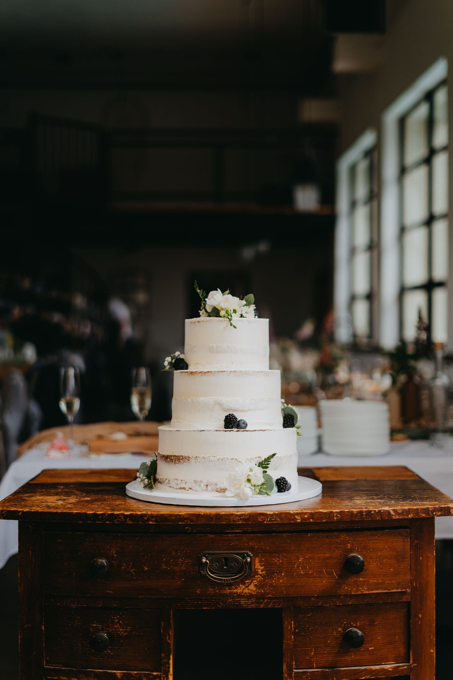 Three-tiered white wedding cake with black berries and floral accents displayed on vintage wooden table by sunlit windows.