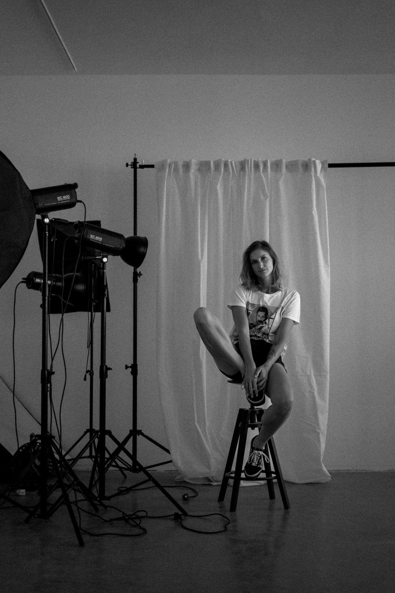 Black and white photo of a photo studio setup with lighting equipment, backdrop, and stool against a white curtain.