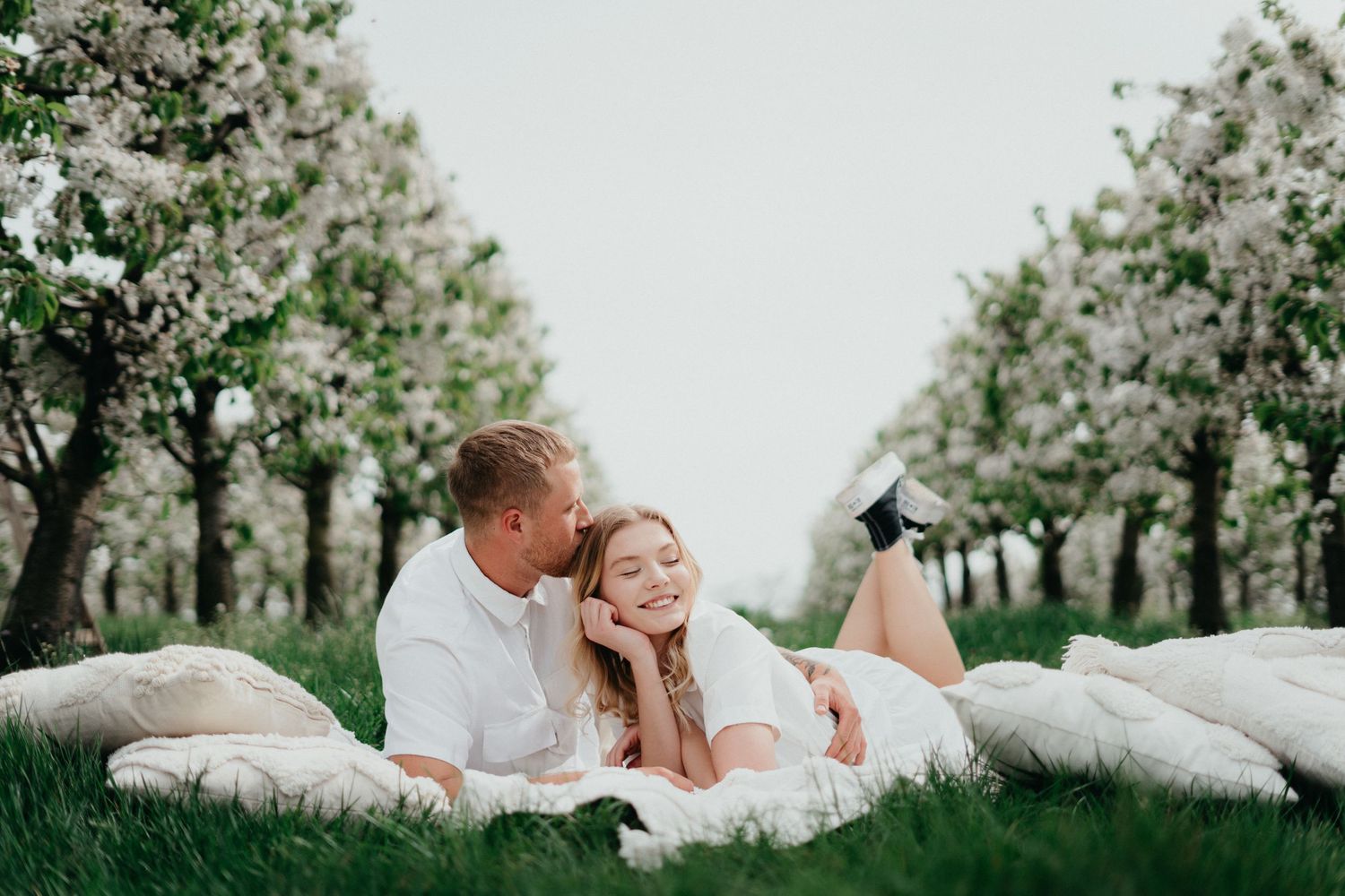 Couple relaxing on white blankets in an orchard of blooming trees.