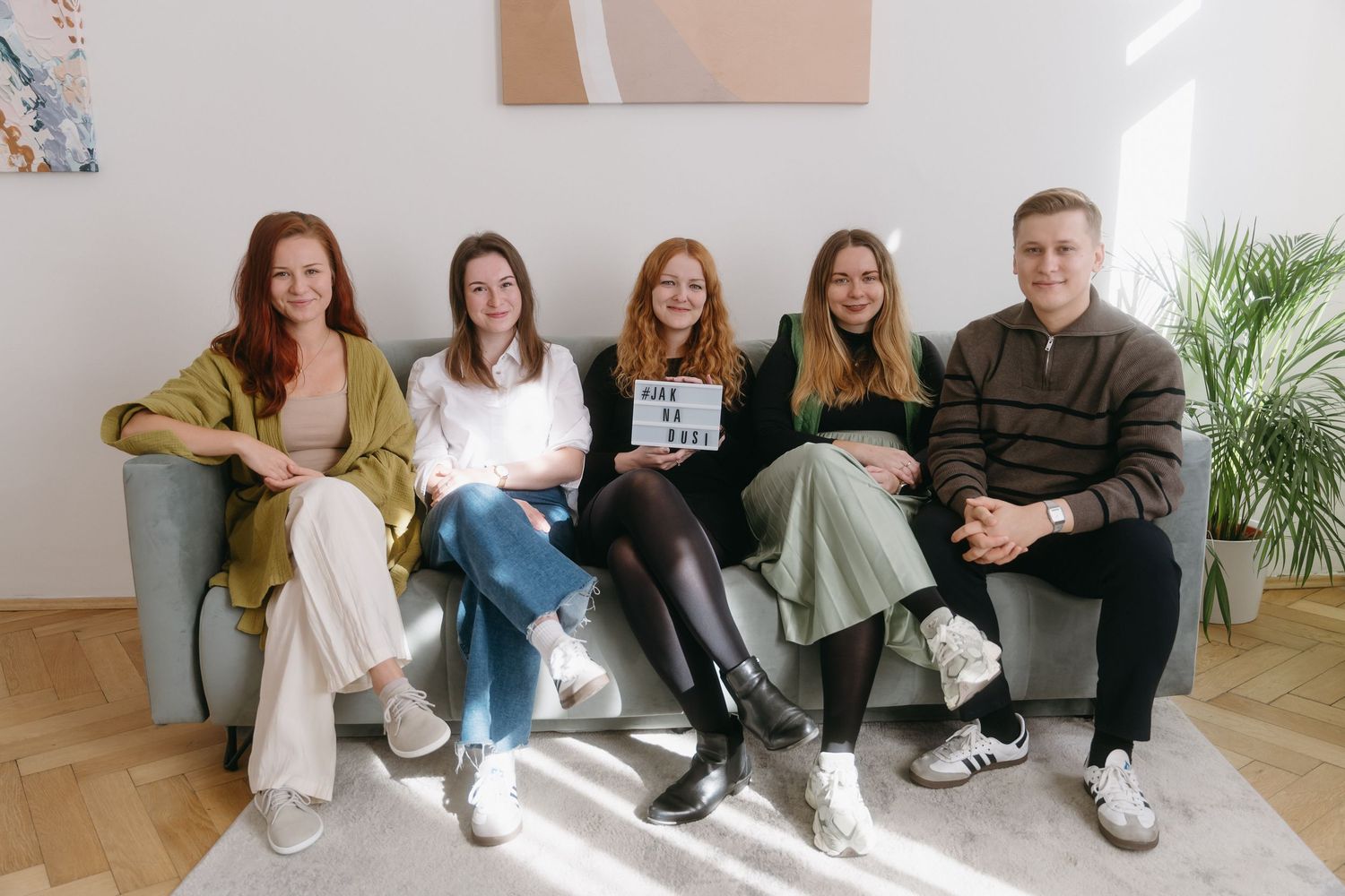 A group of people sitting casually on a white couch in a minimalist room with hardwood floors and plants.