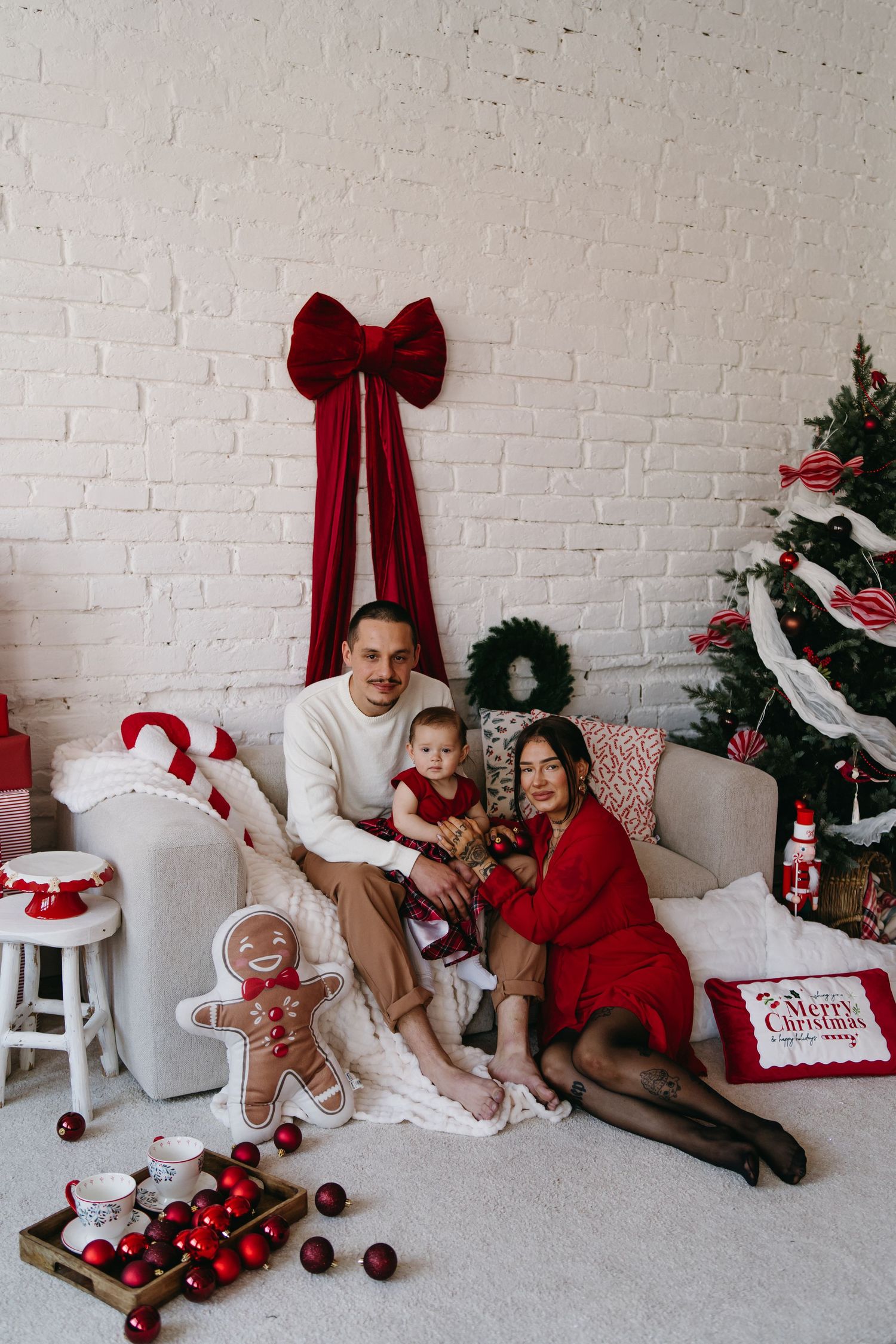 A cozy Christmas scene with a white brick wall, red bow, decorated tree and holiday decorations on the floor.