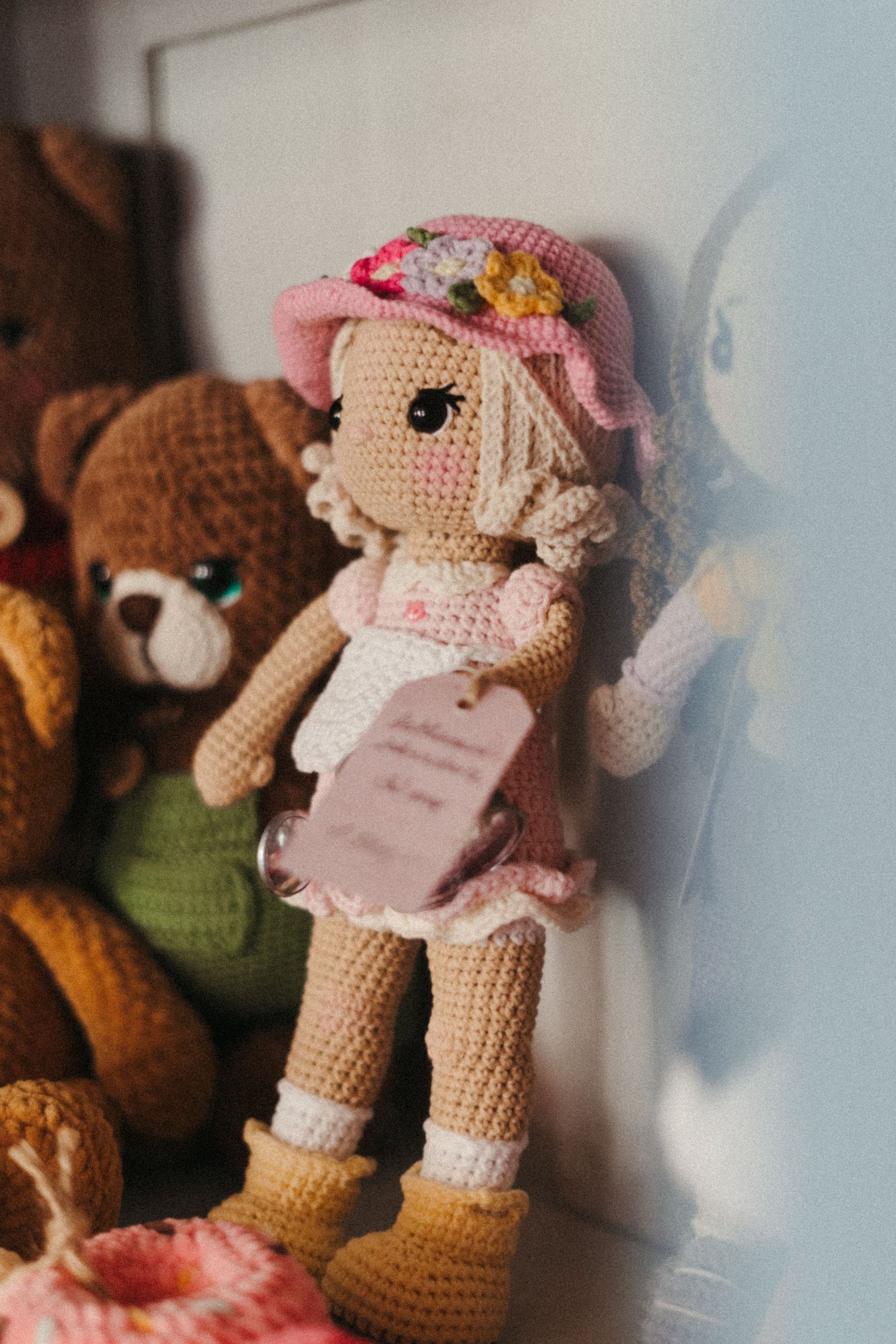 Handmade crochet amigurumi doll wearing pink cap and dress stands next to plush teddy bear on a shelf.
