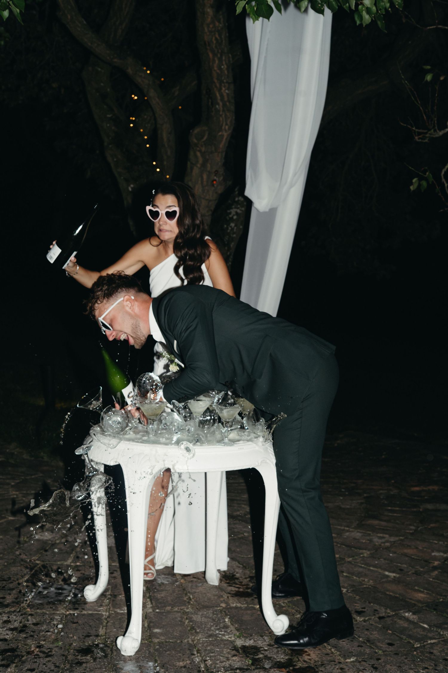 A playful wedding photo showing a couple posing with a decorative white table at an outdoor evening reception.