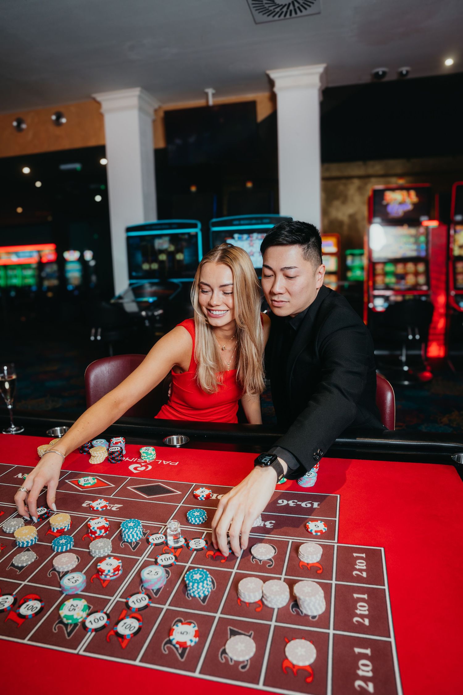 A couple plays roulette at a casino table with red felt and betting chips in a dimly lit gaming room.