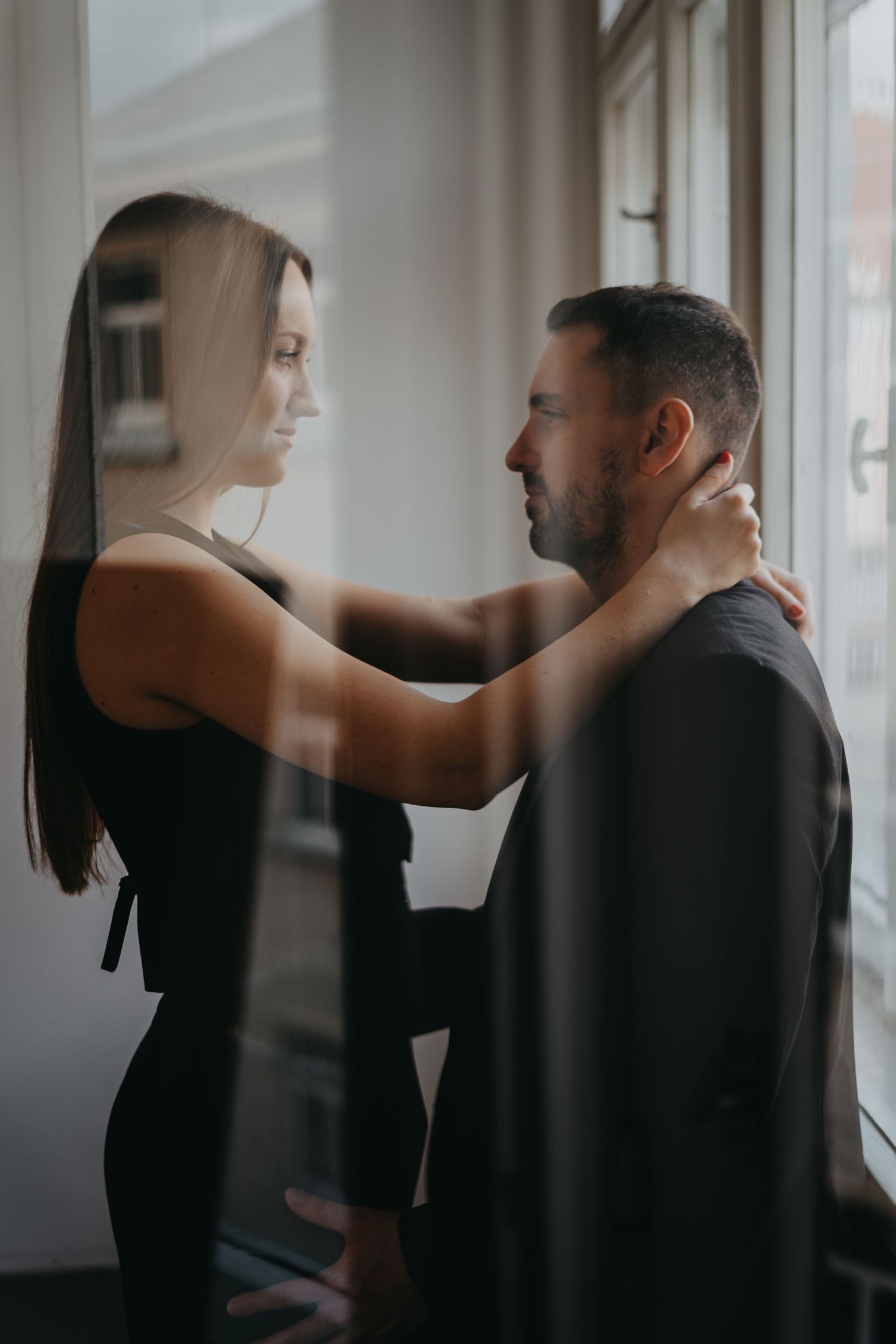 A couple in black clothing embrace tenderly by a window in a romantic indoor setting.