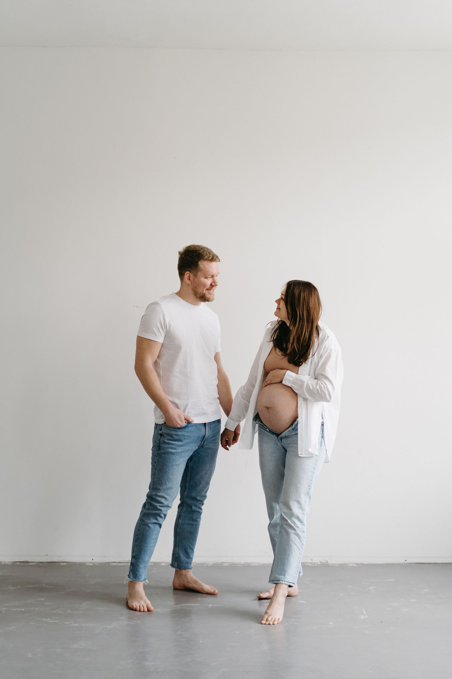 A minimalist maternity photo with a couple in white and denim standing against a plain wall.