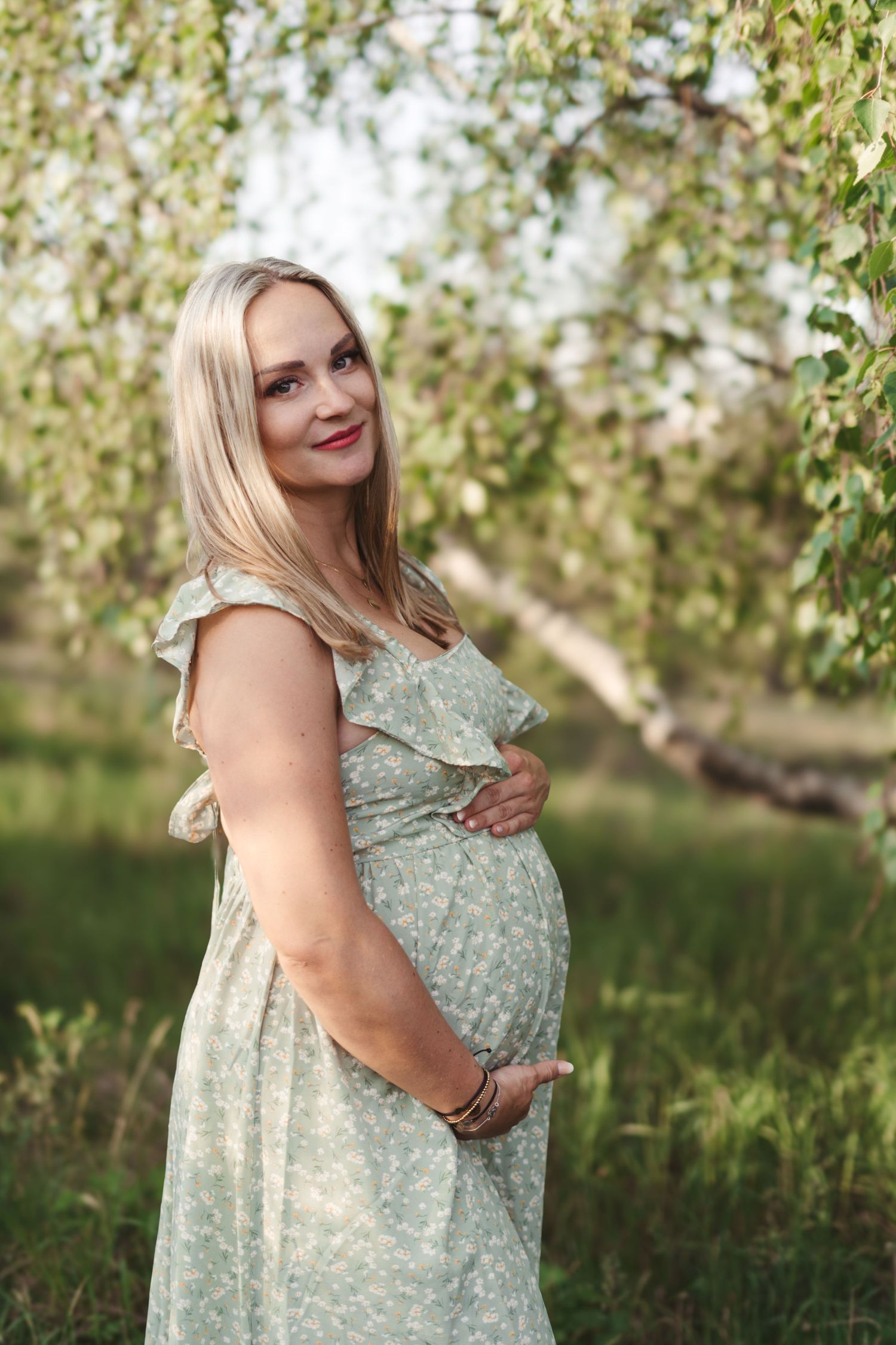 Expectant mother in mint floral dress poses joyfully in outdoor spring setting.