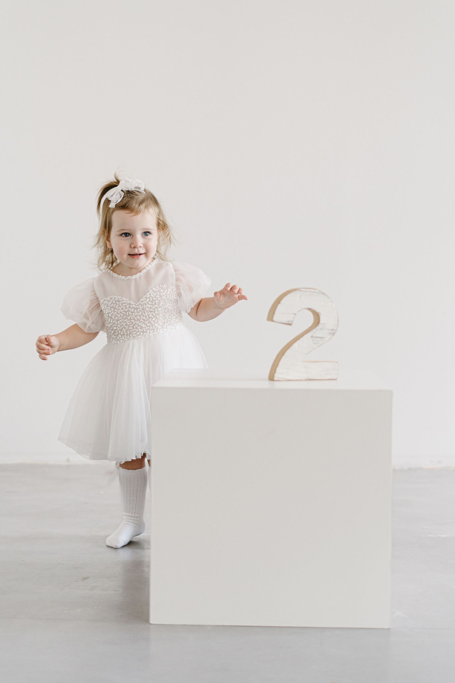 Young child in white dress poses next to number two on white pedestal.
