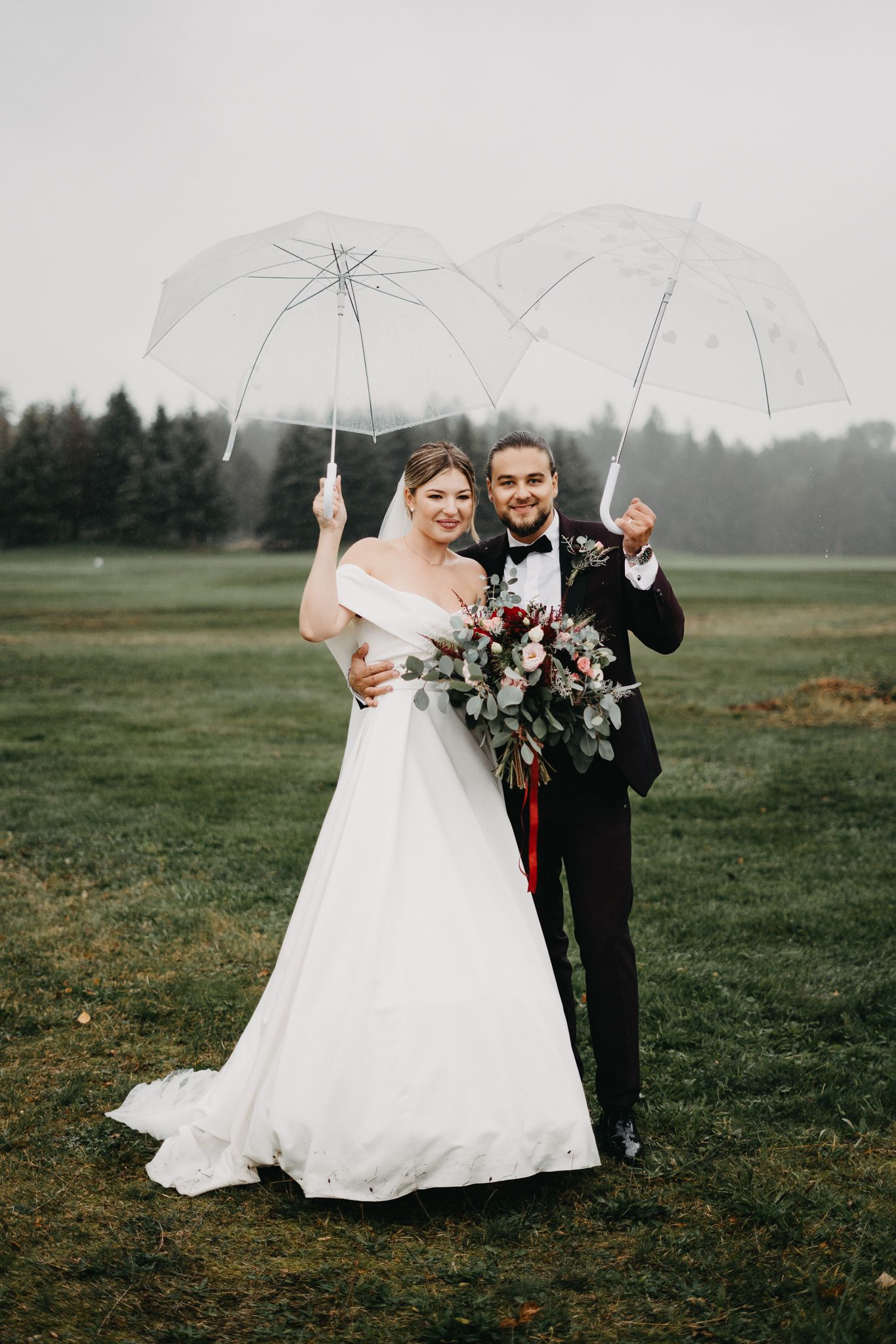 Happy couple holds clear umbrellas together while standing in a misty field with pine trees in the background.