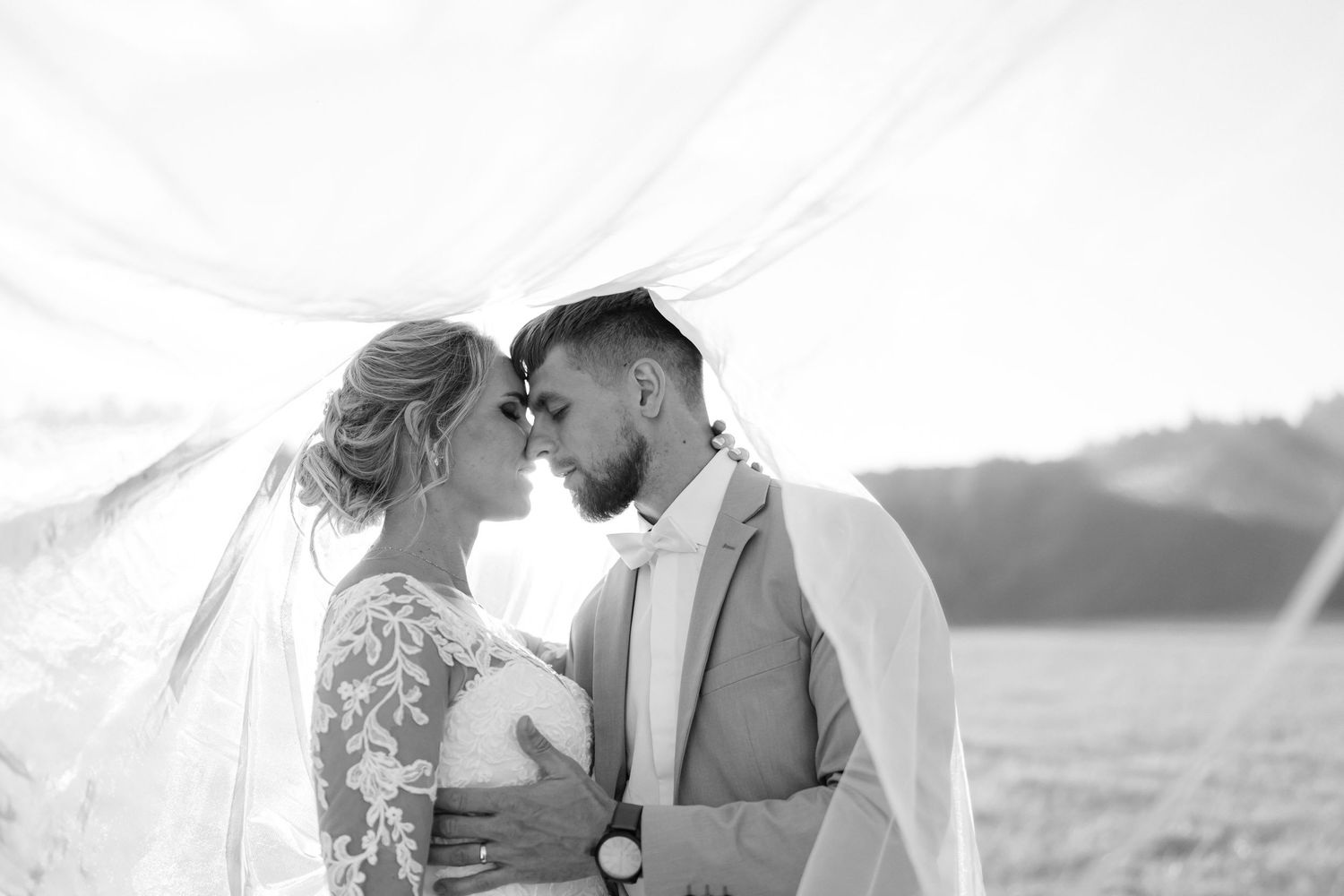 A romantic black and white wedding photo of couple kissing under a flowing veil in a mountain field.