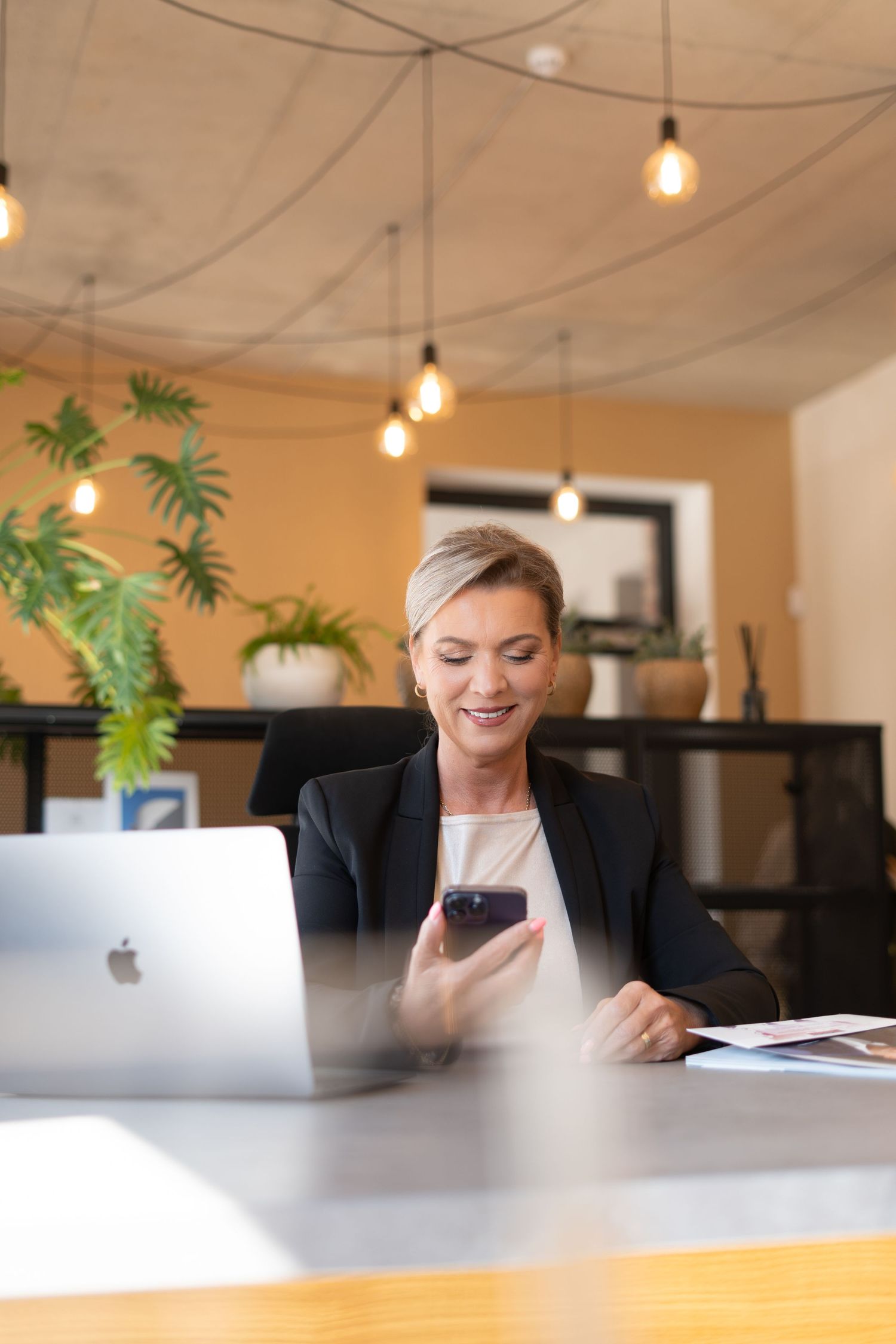 Professional businesswoman in modern office working on MacBook laptop and smartphone under pendant lighting.