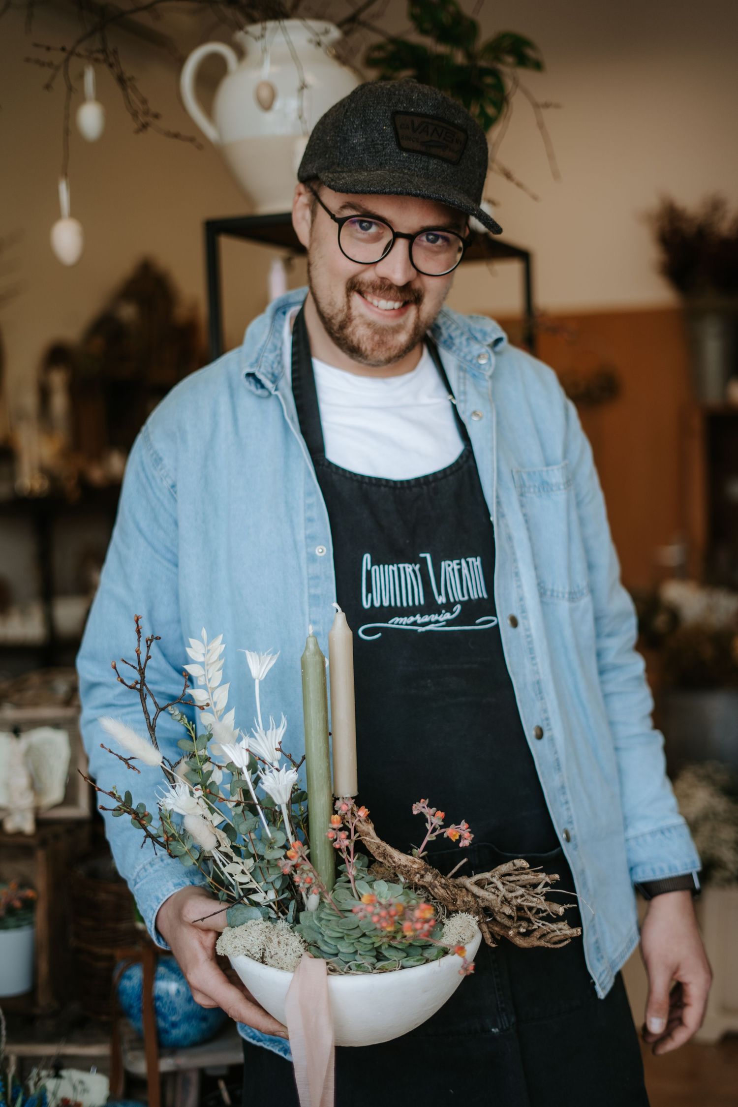 A small business employee wearing a baseball cap and denim jacket holds a decorative floral arrangement in a white bowl.