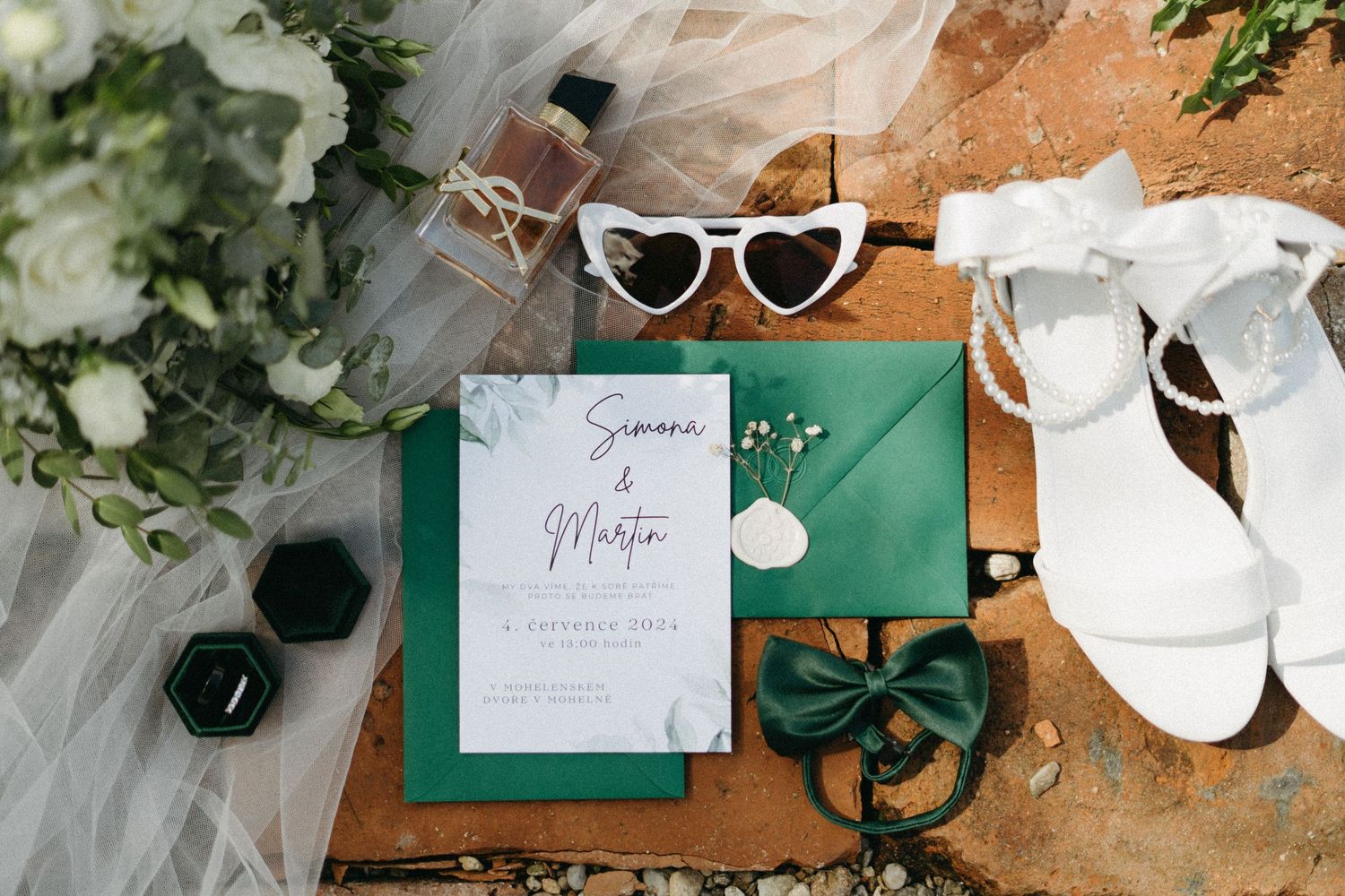 Wedding accessories flatlay with emerald green invitation, heart-shaped sunglasses, veil and white bridal shoes on wooden surface.