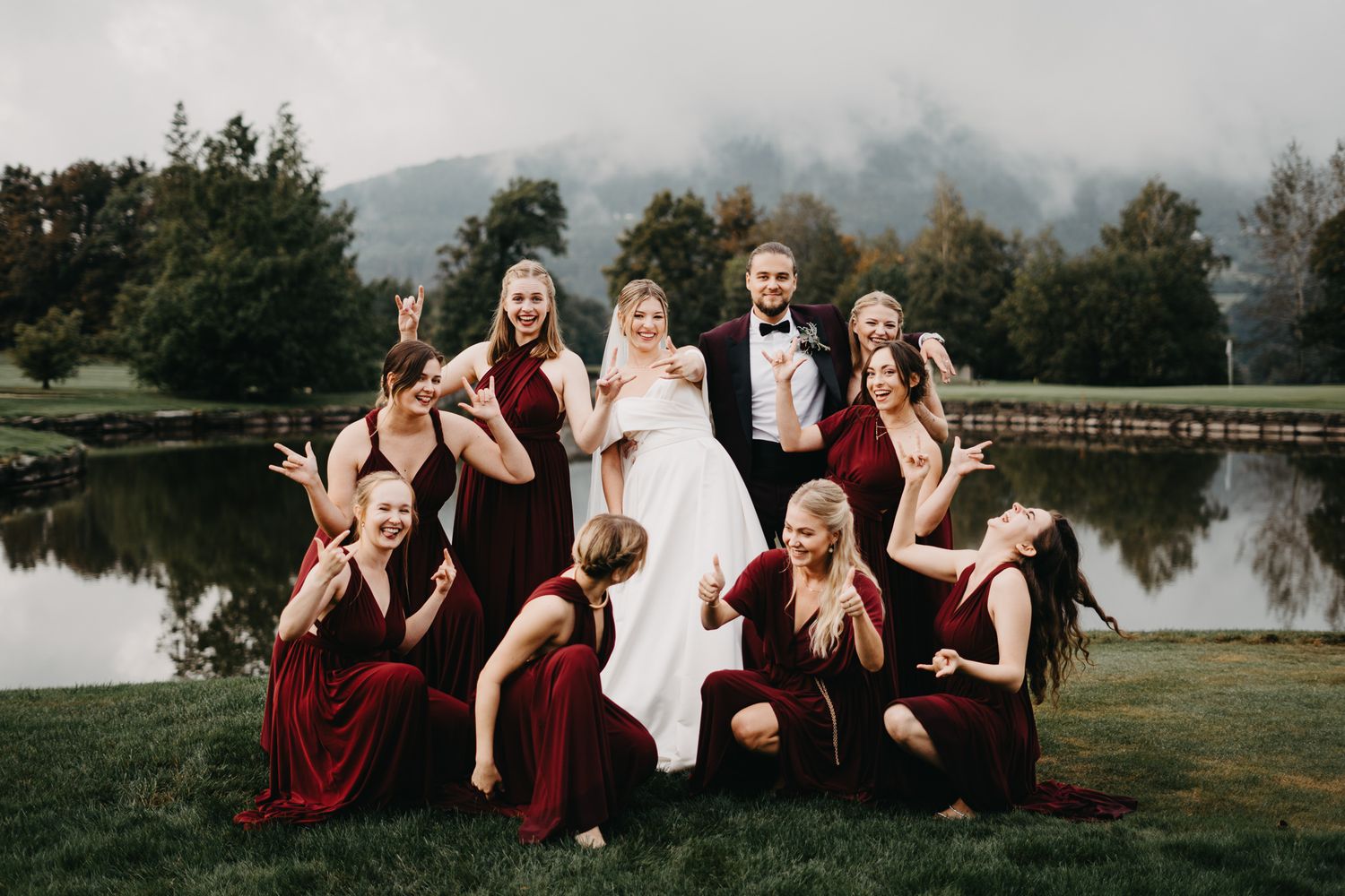 Wedding party poses together on grass near a serene lake with mountains in the background.