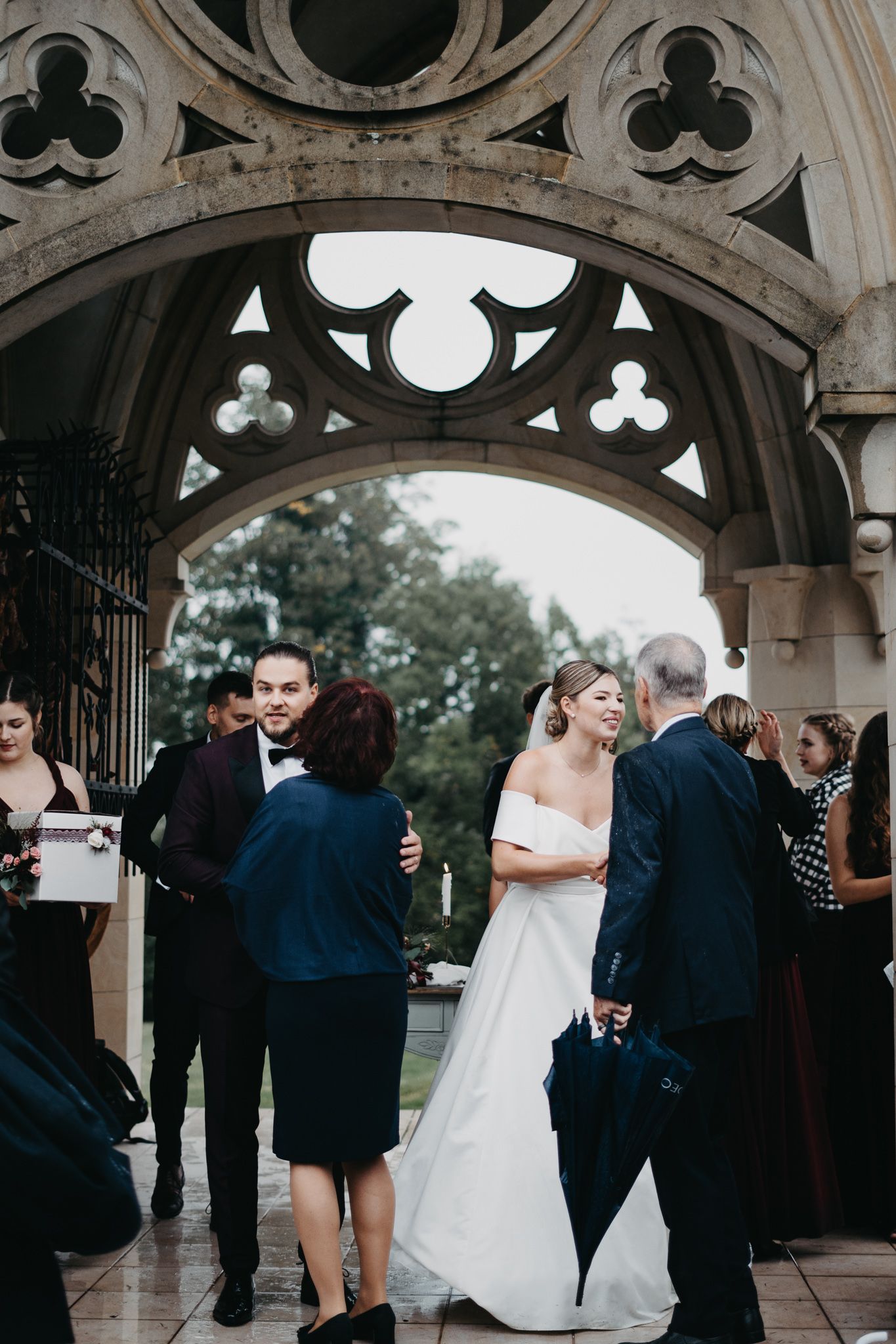 Wedding guests gather under a Gothic stone archway with decorative trefoil details during an outdoor ceremony.