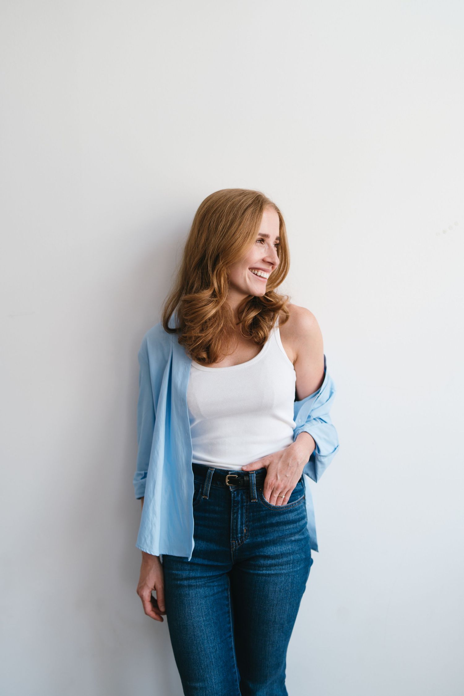 Smiling redhead woman in white tank top, blue shirt, and jeans leaning against a white wall.