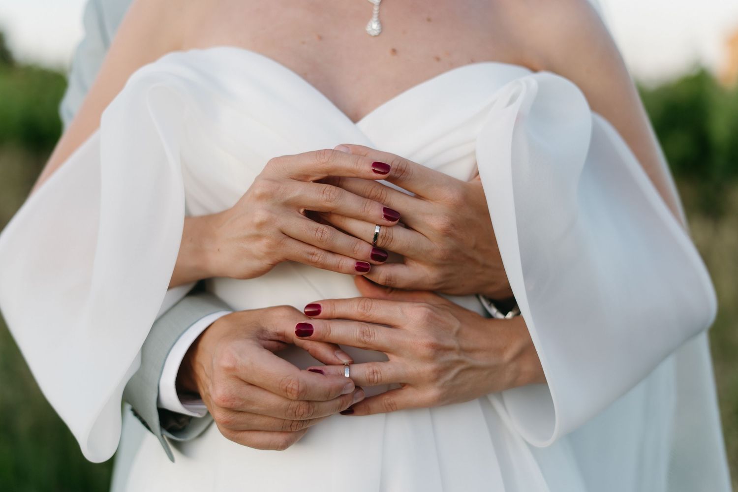 Close-up photo of hands with red manicured nails resting on white wedding dress fabric.