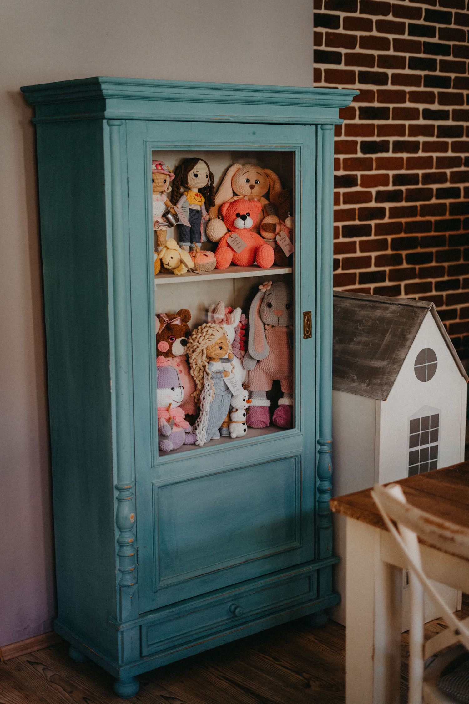 A teal-colored vintage cabinet displays a collection of plush stuffed animals and toys against a brick wall interior.