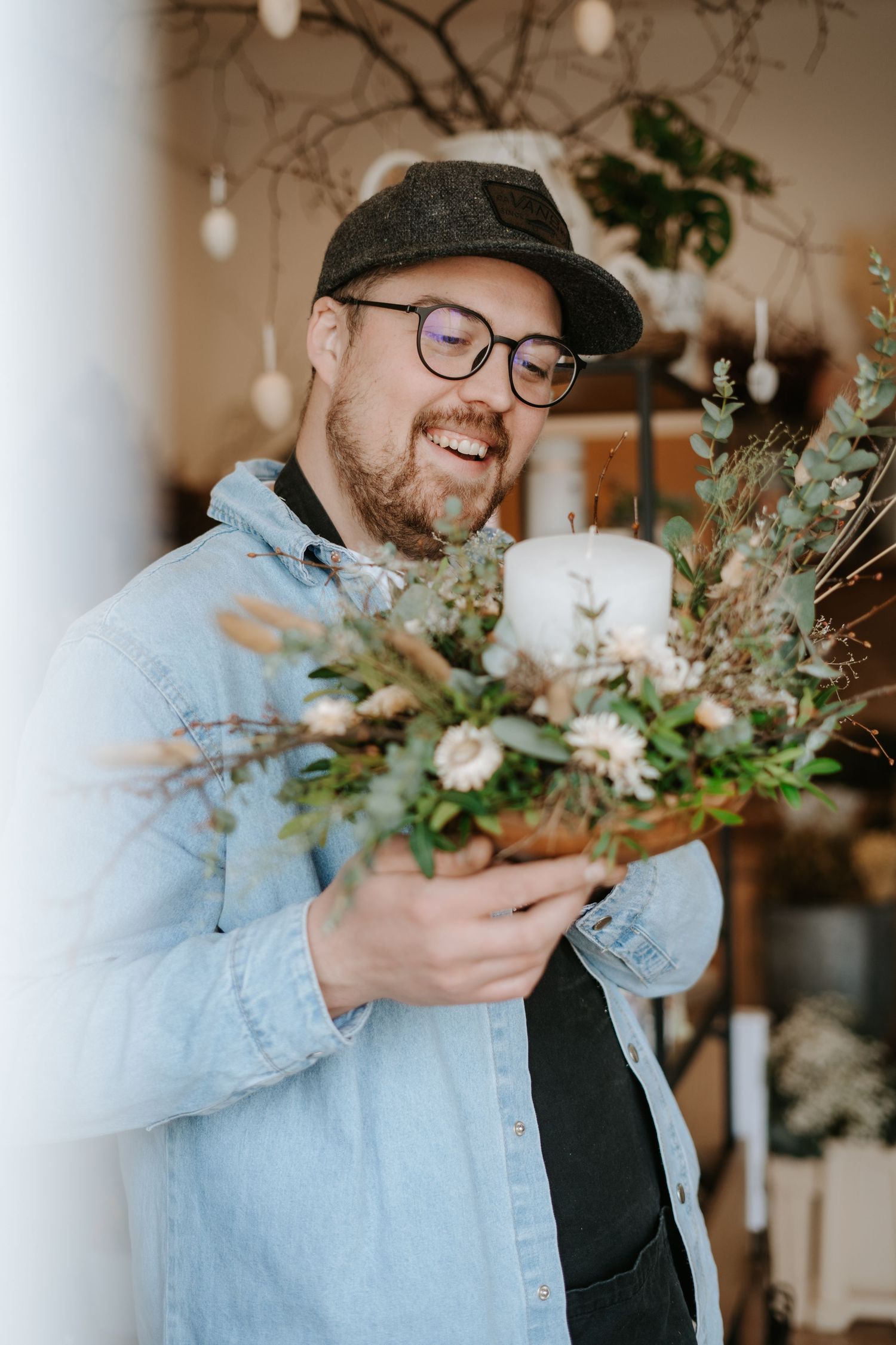 Person in denim jacket and glasses smiles while holding white floral arrangements in vintage-style indoor setting.