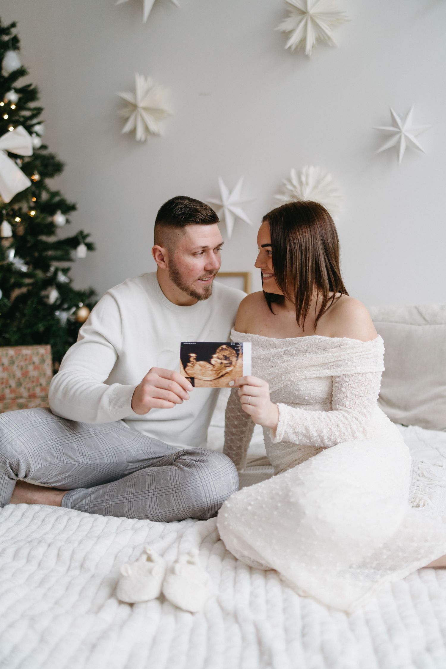 Happy couple sitting on bed holding ultrasound photos near Christmas tree, announcing pregnancy during holidays.
