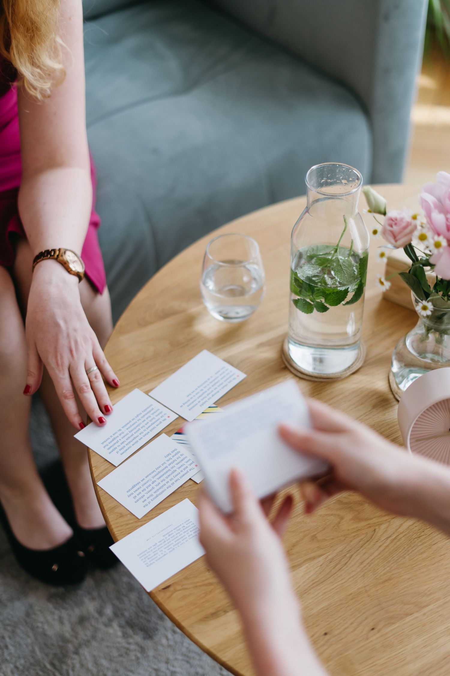 Two people discussing with papers on a wooden coffee table next to a glass water bottle with fresh mint and flowers.