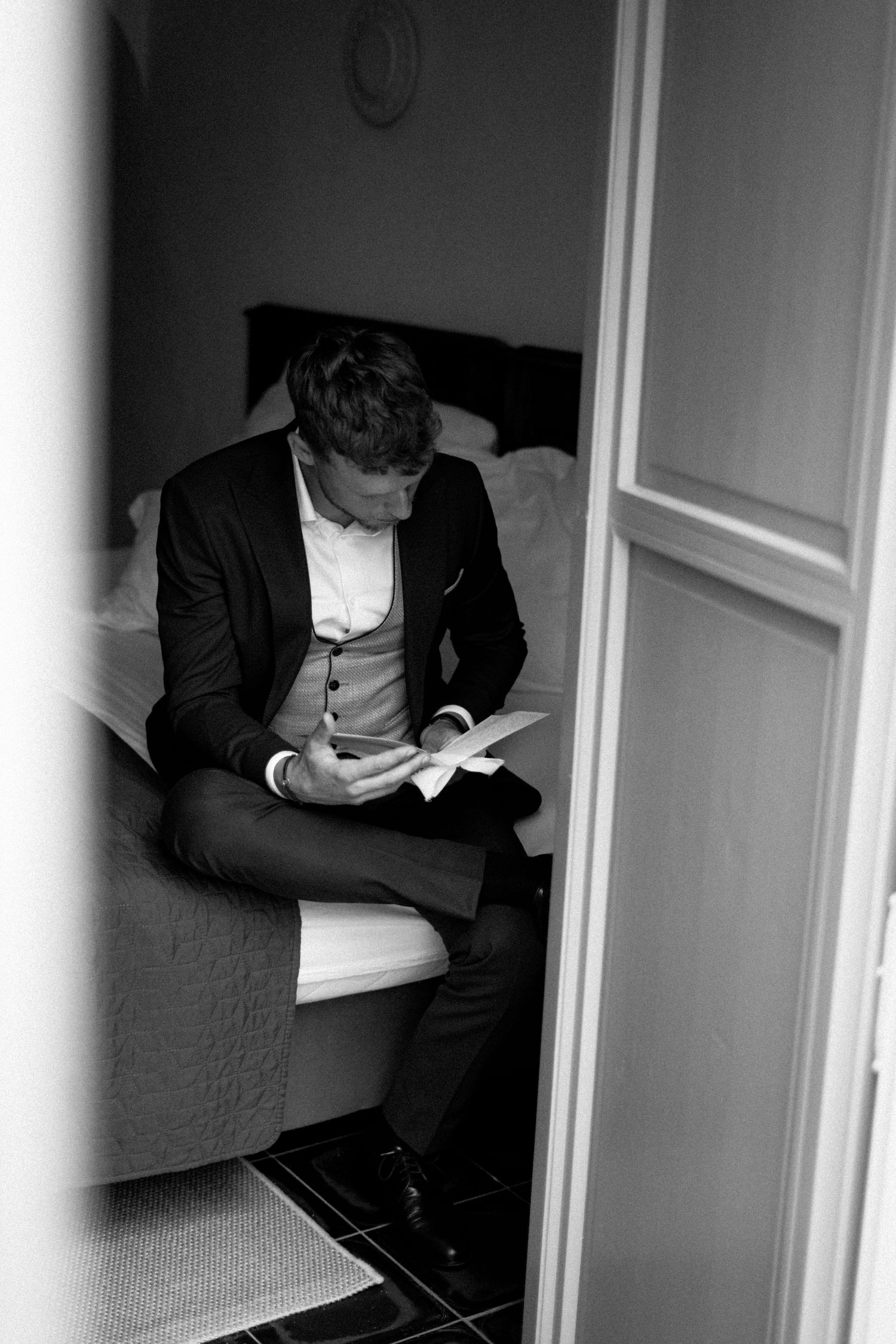 Person in formal black suit sitting in corner of white room looking downward.